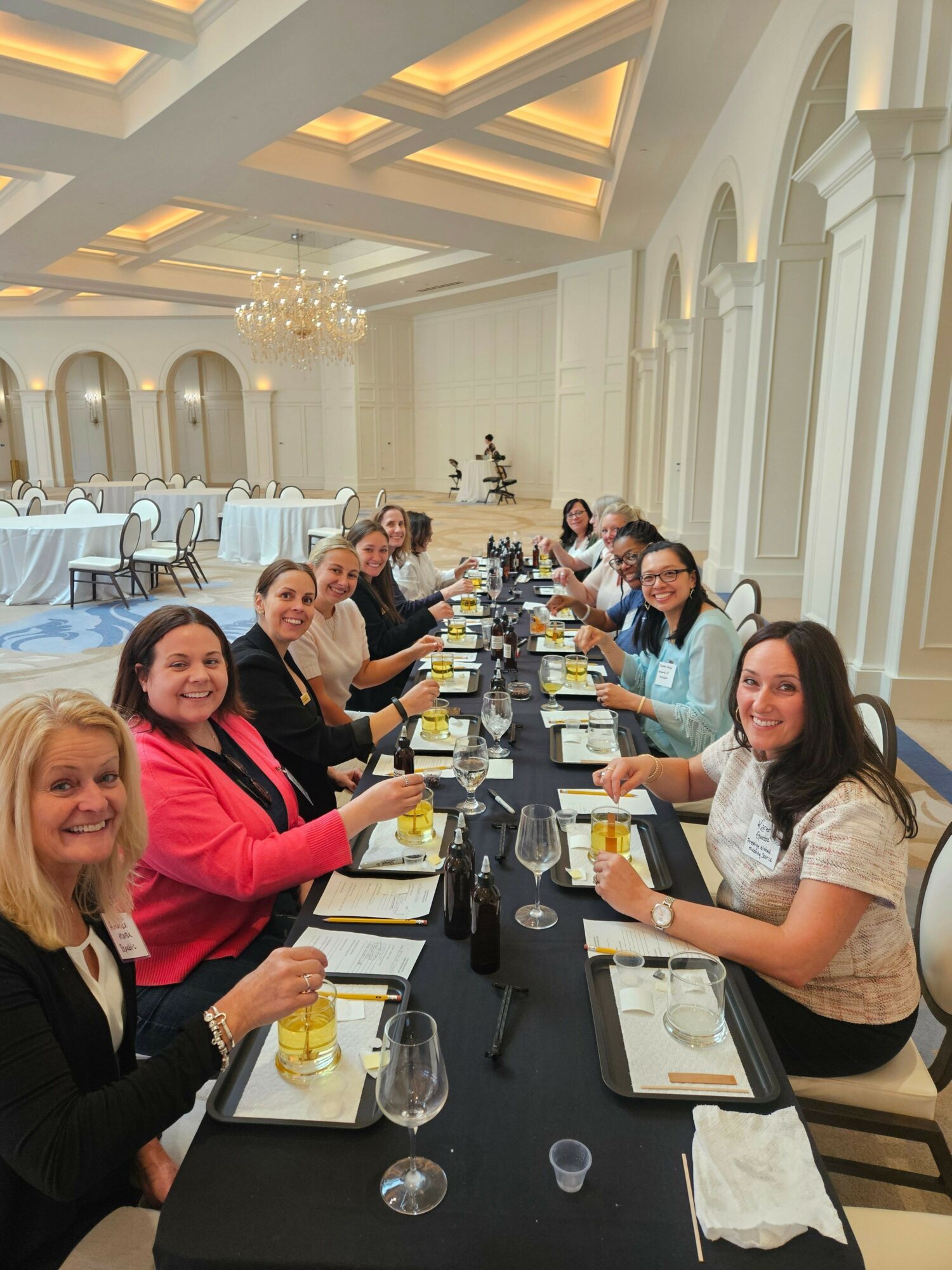 People sitting at a long table with drinks and food in a well-lit, elegant room with high ceilings and chandeliers.