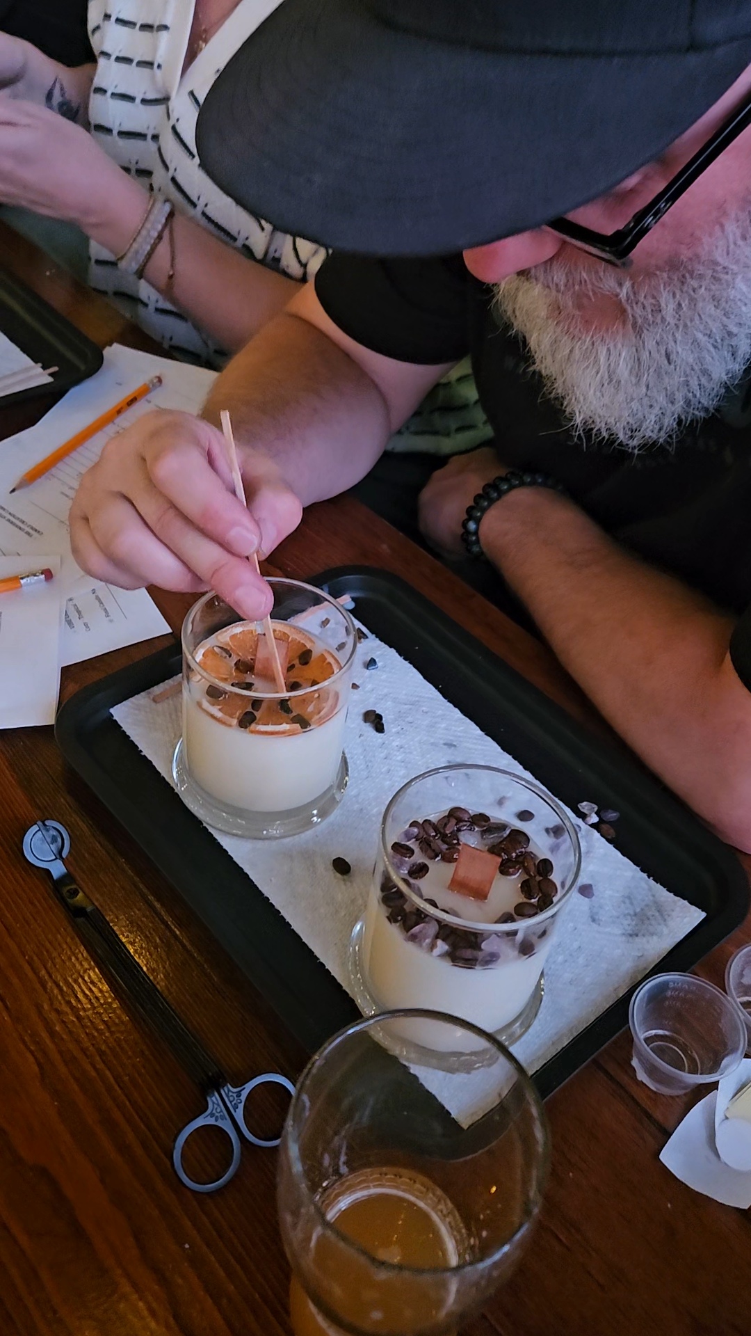 Man with a beard and glasses, wearing a hat, is decorating desserts with a spoon. Two desserts are on a tray.