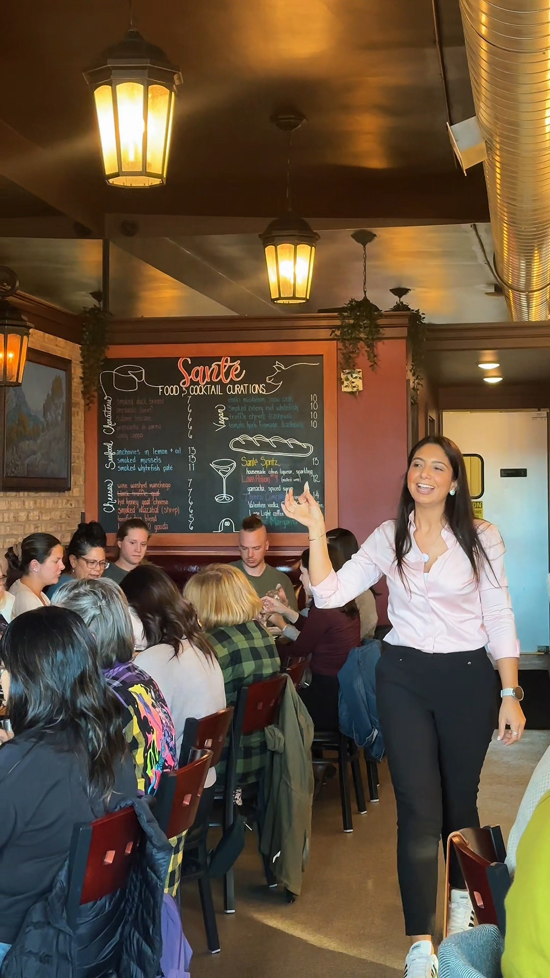 Woman standing and pointing at a chalkboard menu in a restaurant with seated customers and hanging lights.