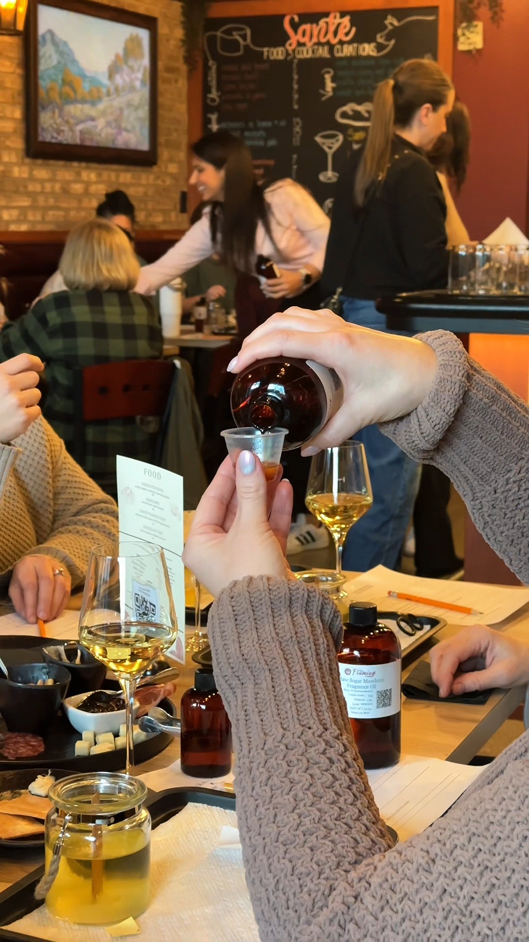 Person pouring dark liquid into a small glass at a busy cafe with people and a menu board in background.