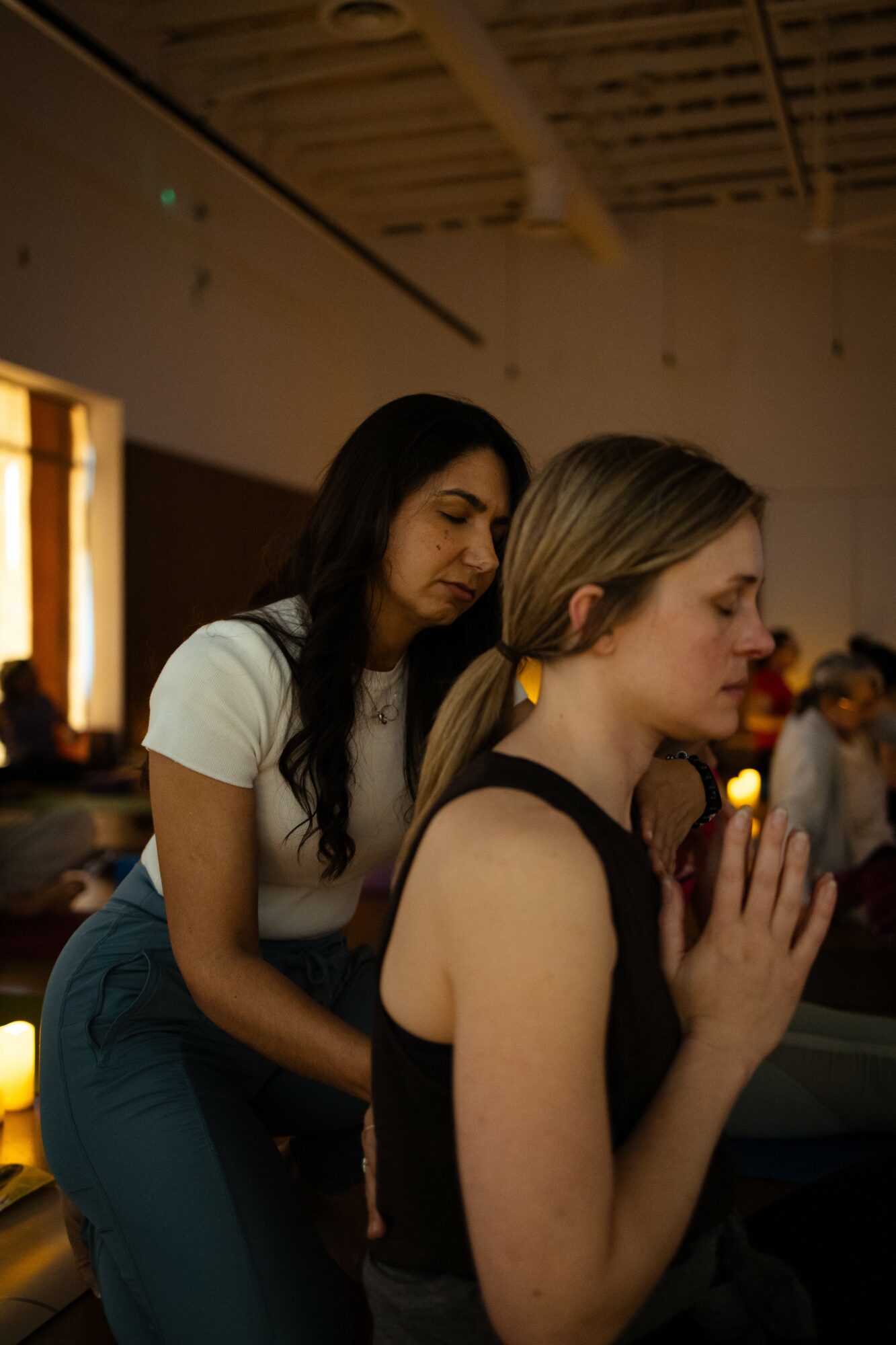 Two women in a dimly lit room, one with hands clasped, eyes closed, and the other with eyes closed, hands together, in prayer.