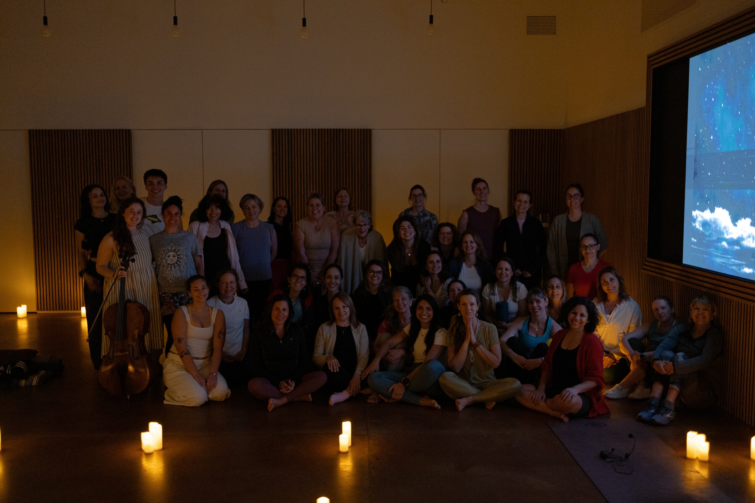 Group of people gathered in a dimly lit room with candles, some sitting and some standing, with a large screen on the right.