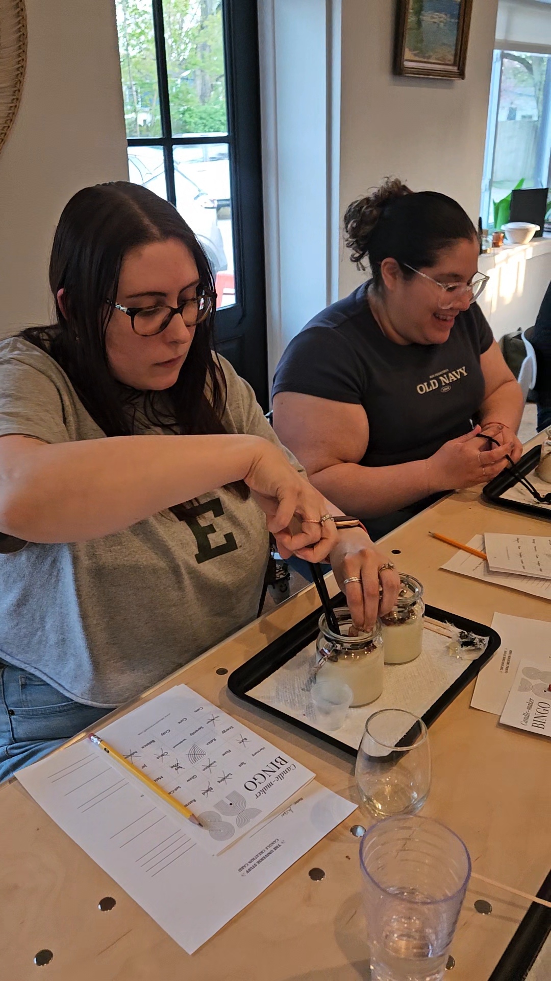 Two women sit at a table, pouring liquid into glasses, with papers and a tray on the table.