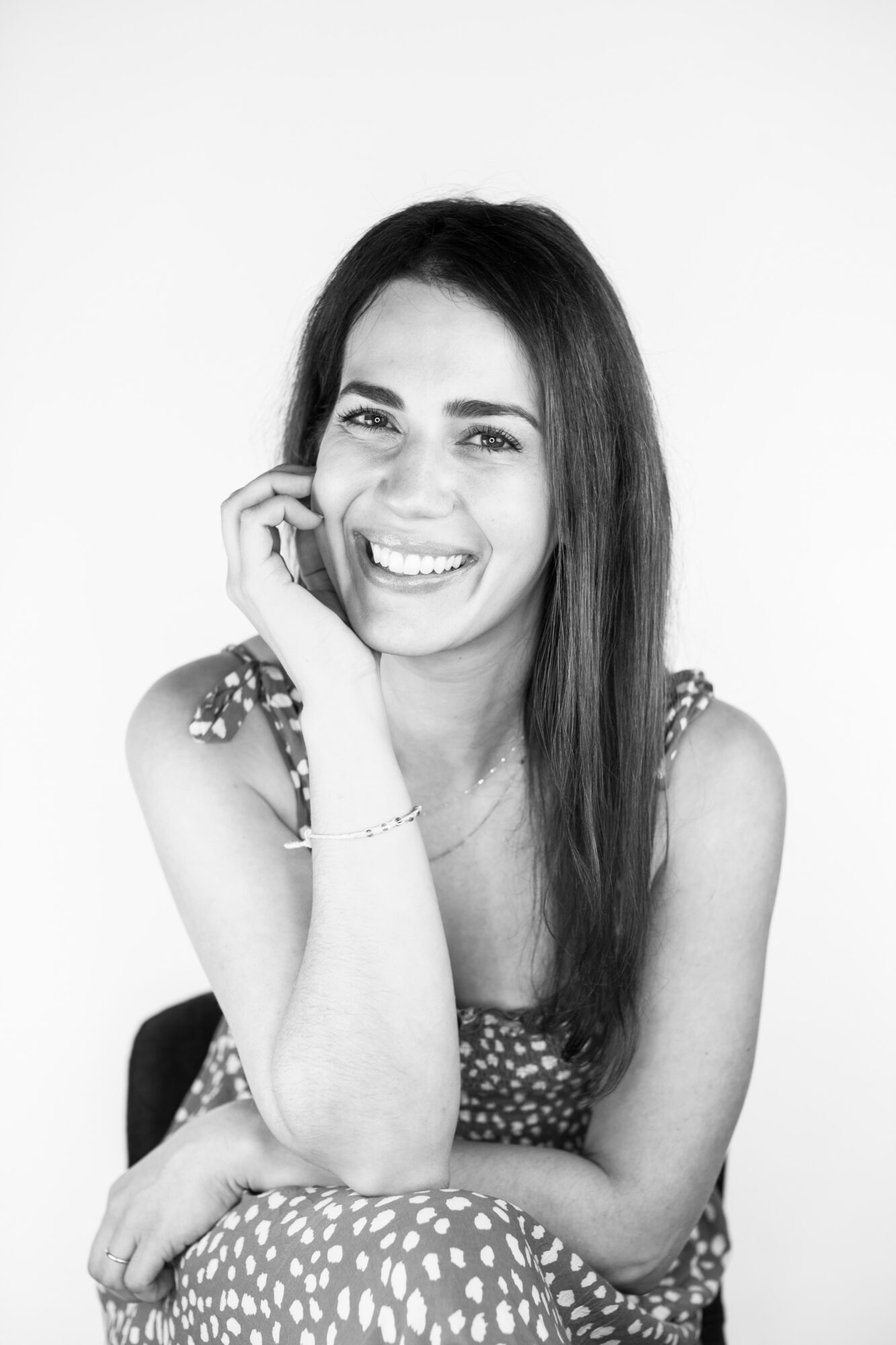 Smiling woman with long hair, resting her chin on her hand, wearing a patterned dress, in black and white.