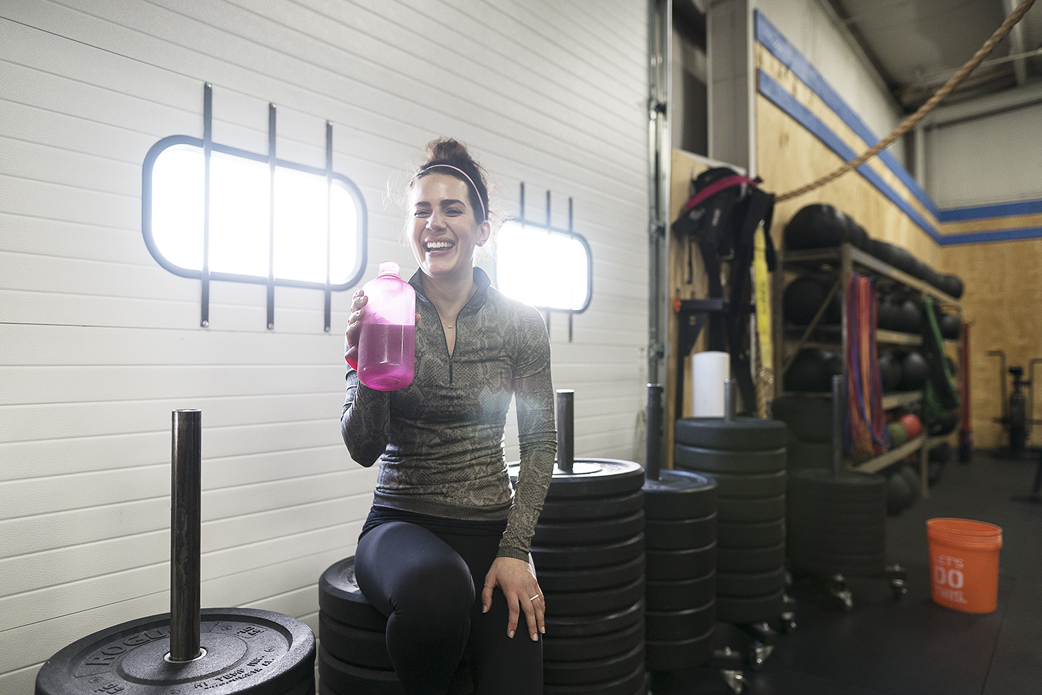 Woman in gym holding pink water bottle, smiling, standing near stacked weights and gym equipment, illuminated by bright light.