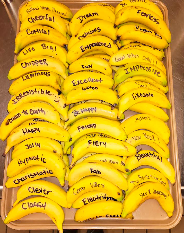 Bananas with handwritten positive words and phrases on each peel, arranged in rows inside a container.