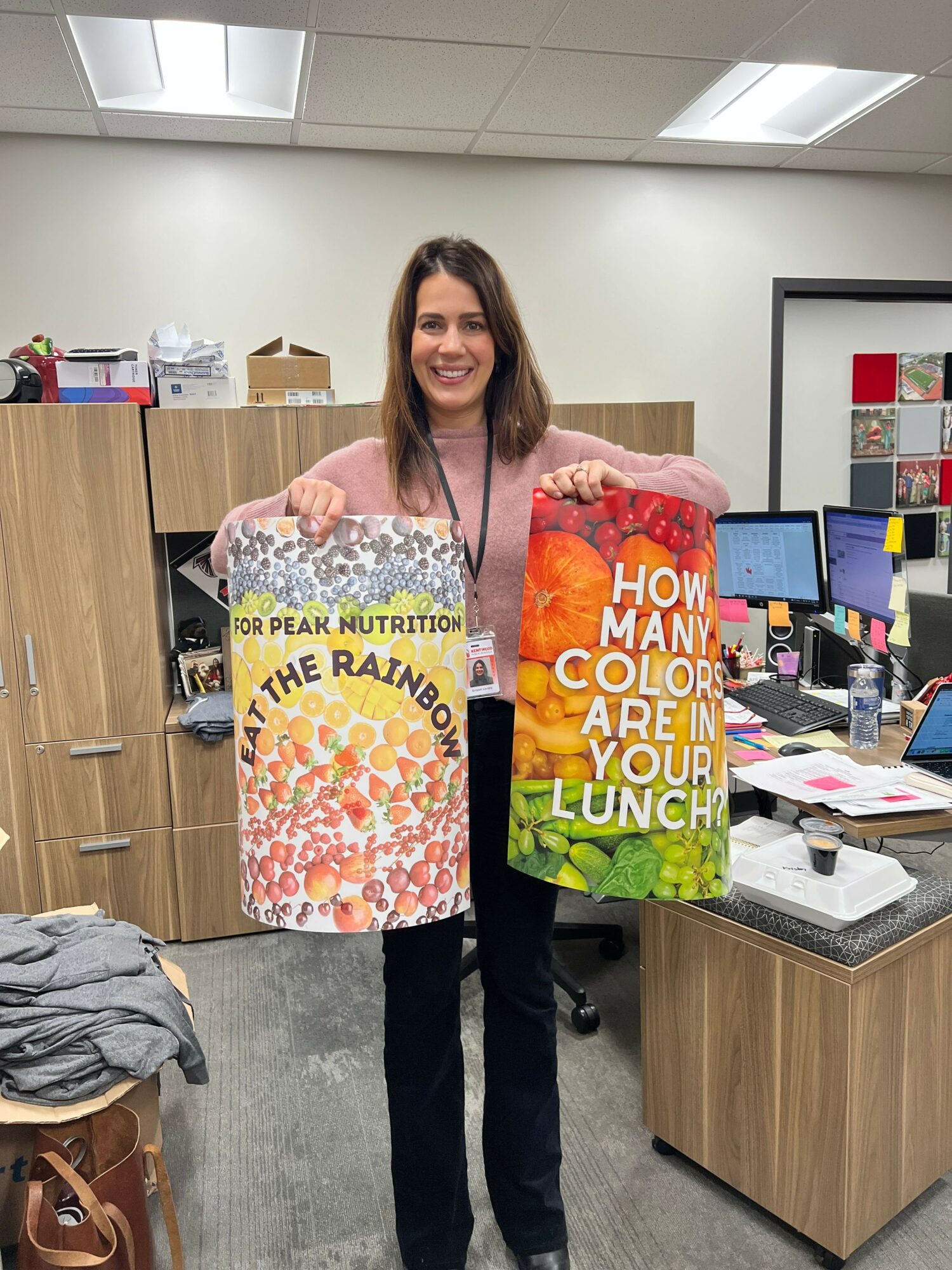 Woman holding a colorful box with text, standing in an office with a desk and shelves behind her.