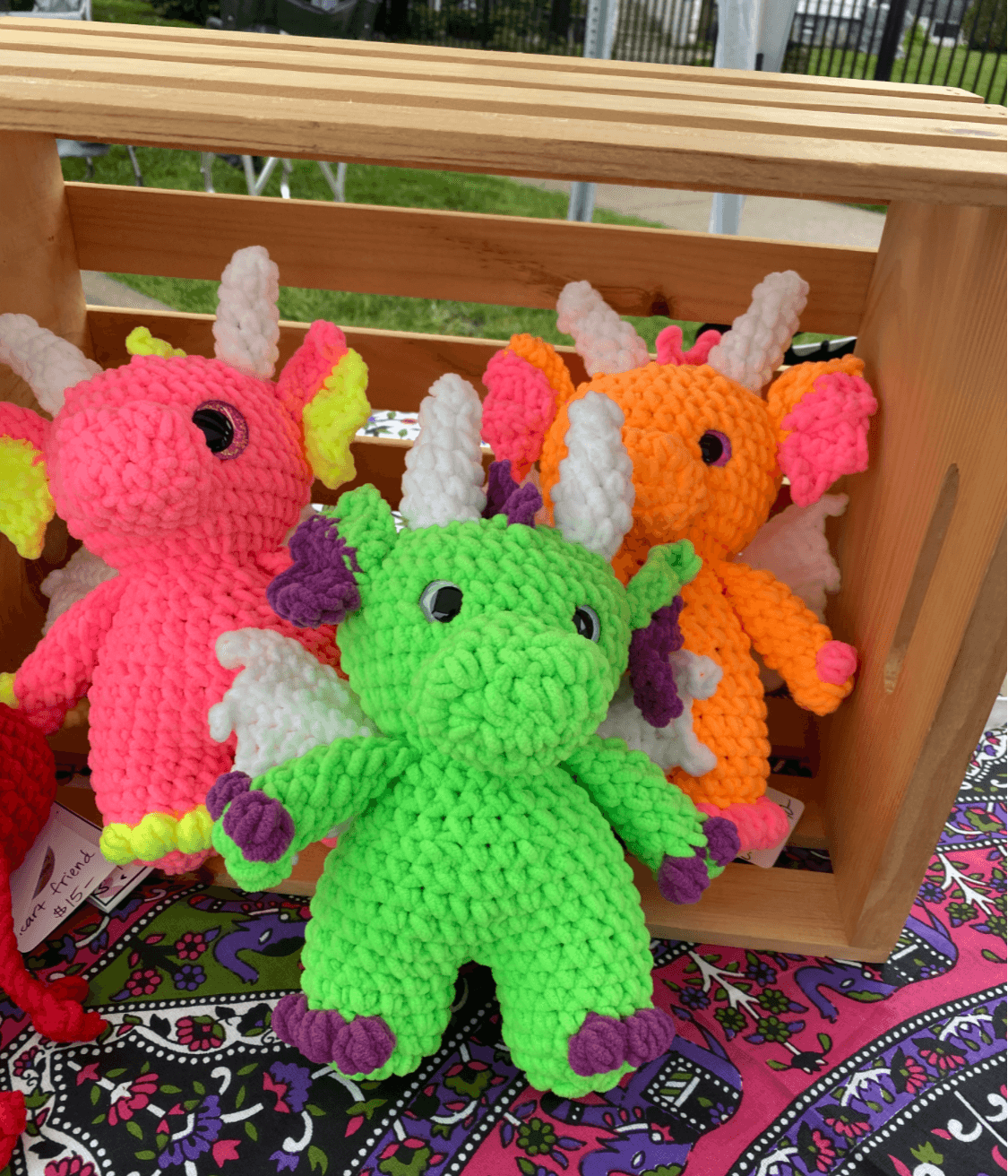 Four colorful crocheted dragon plush toys inside a wooden box on a patterned fabric surface.