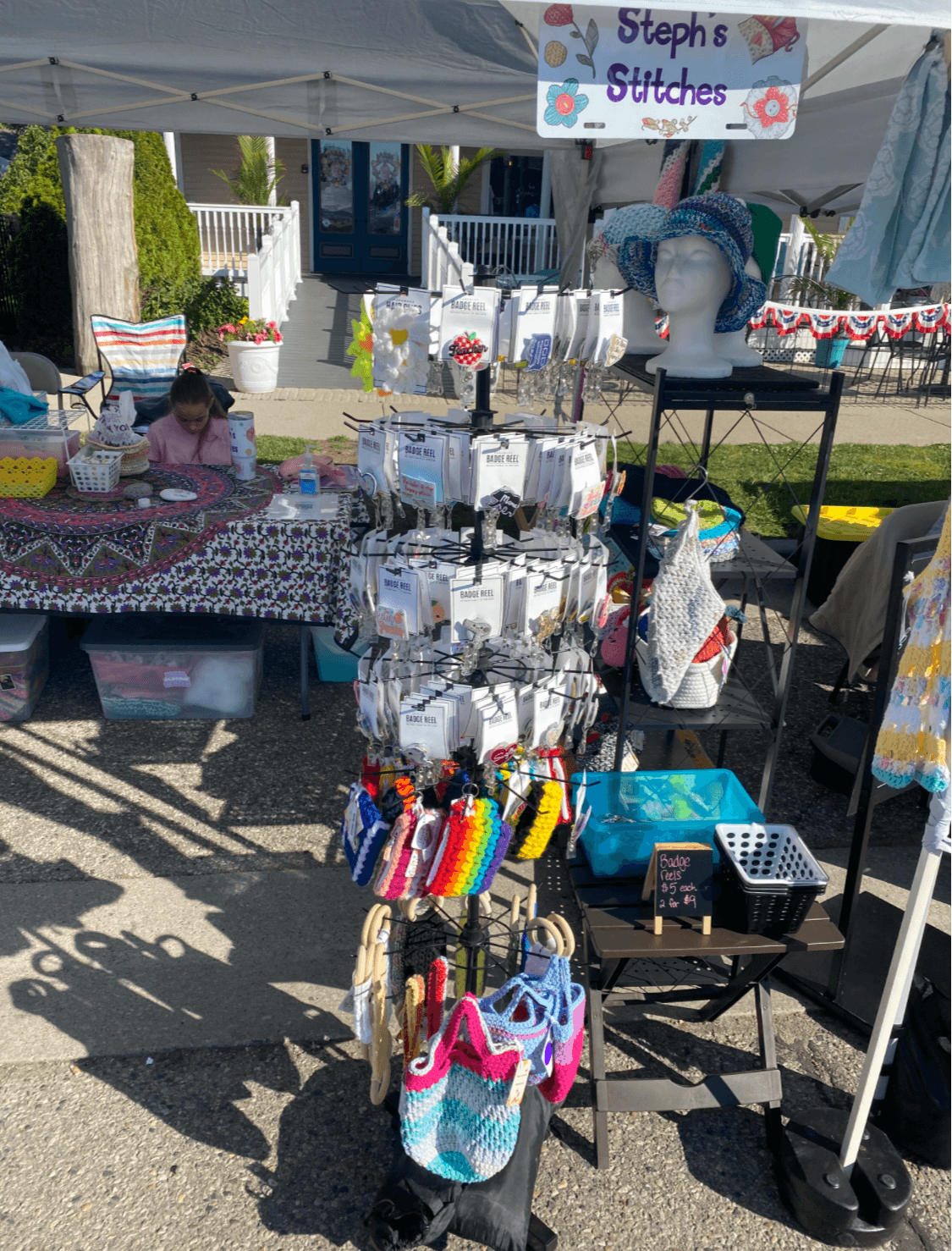 Display stand with handmade stitched items at outdoor market, with tents, tables, and people in background.