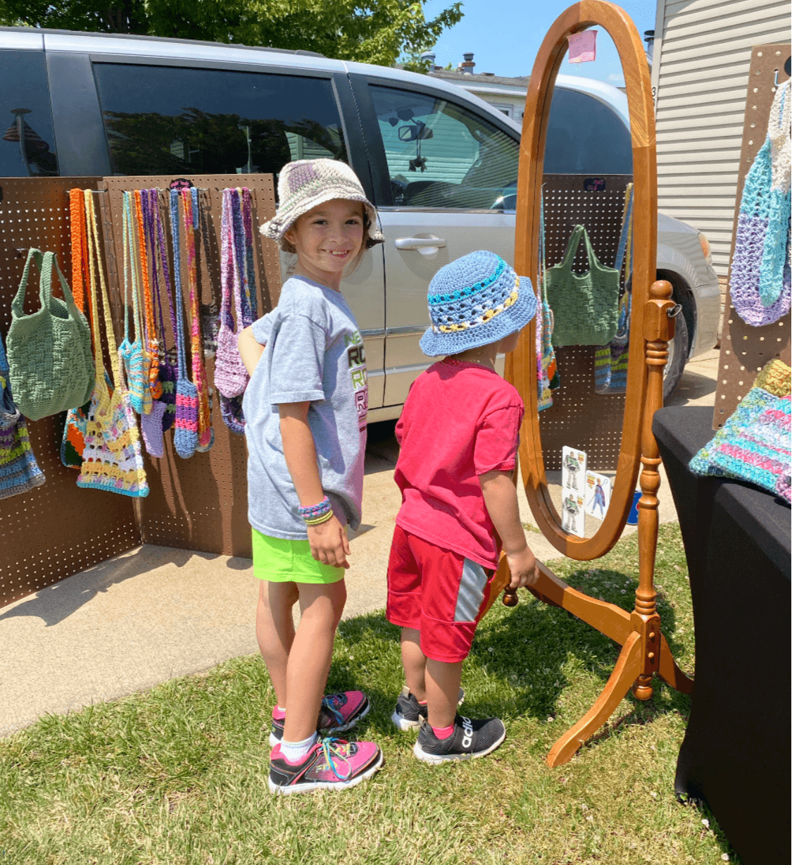 Two children browsing colorful knitted items at an outdoor market stall, with a vehicle and greenery in the background.