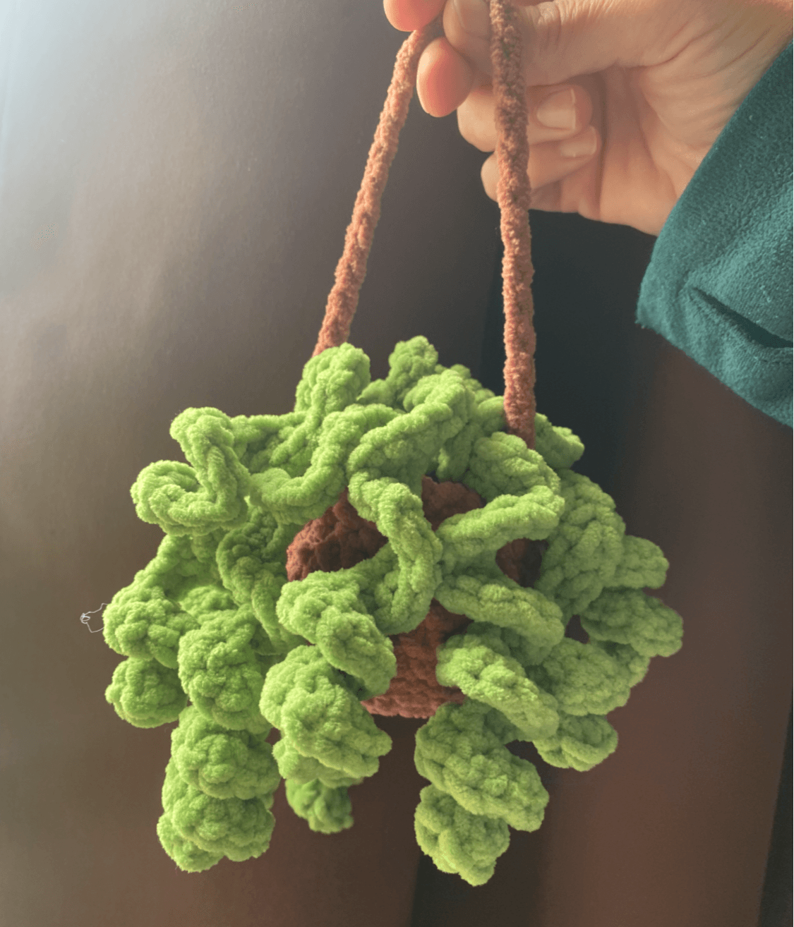 Hand holding a green, fluffy, pom-pom-like object with a brown string handle, against a dark background.
