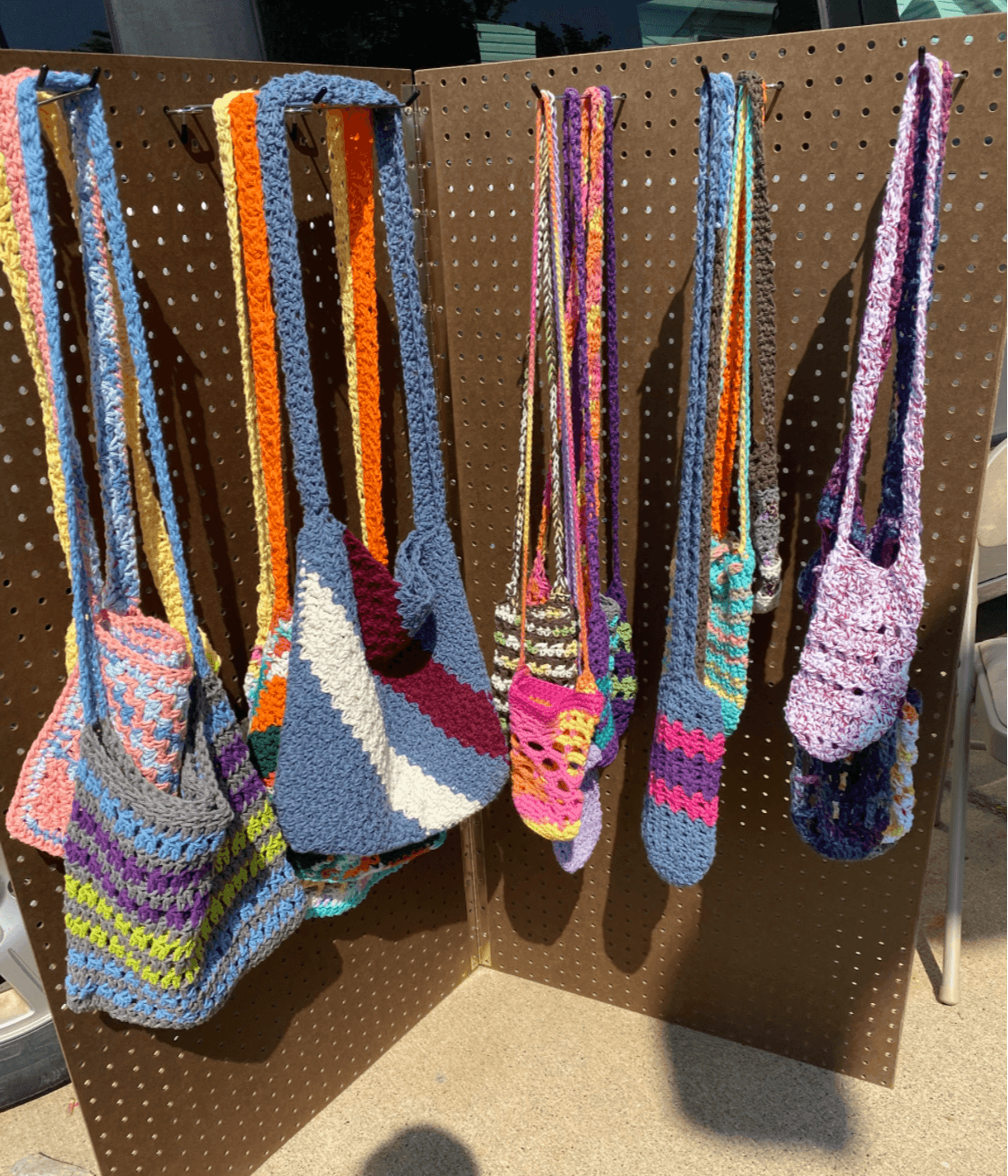 Colorful knitted bags hanging on a display board, with a shadow cast on the ground.