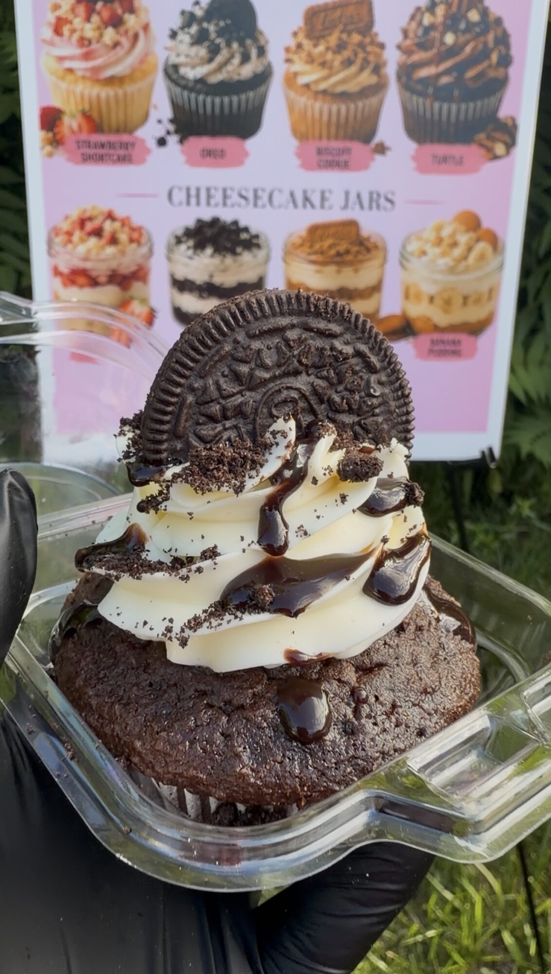 Chocolate cupcake topped with white frosting, chocolate drizzle, and cookie crumbs, in a clear plastic container.