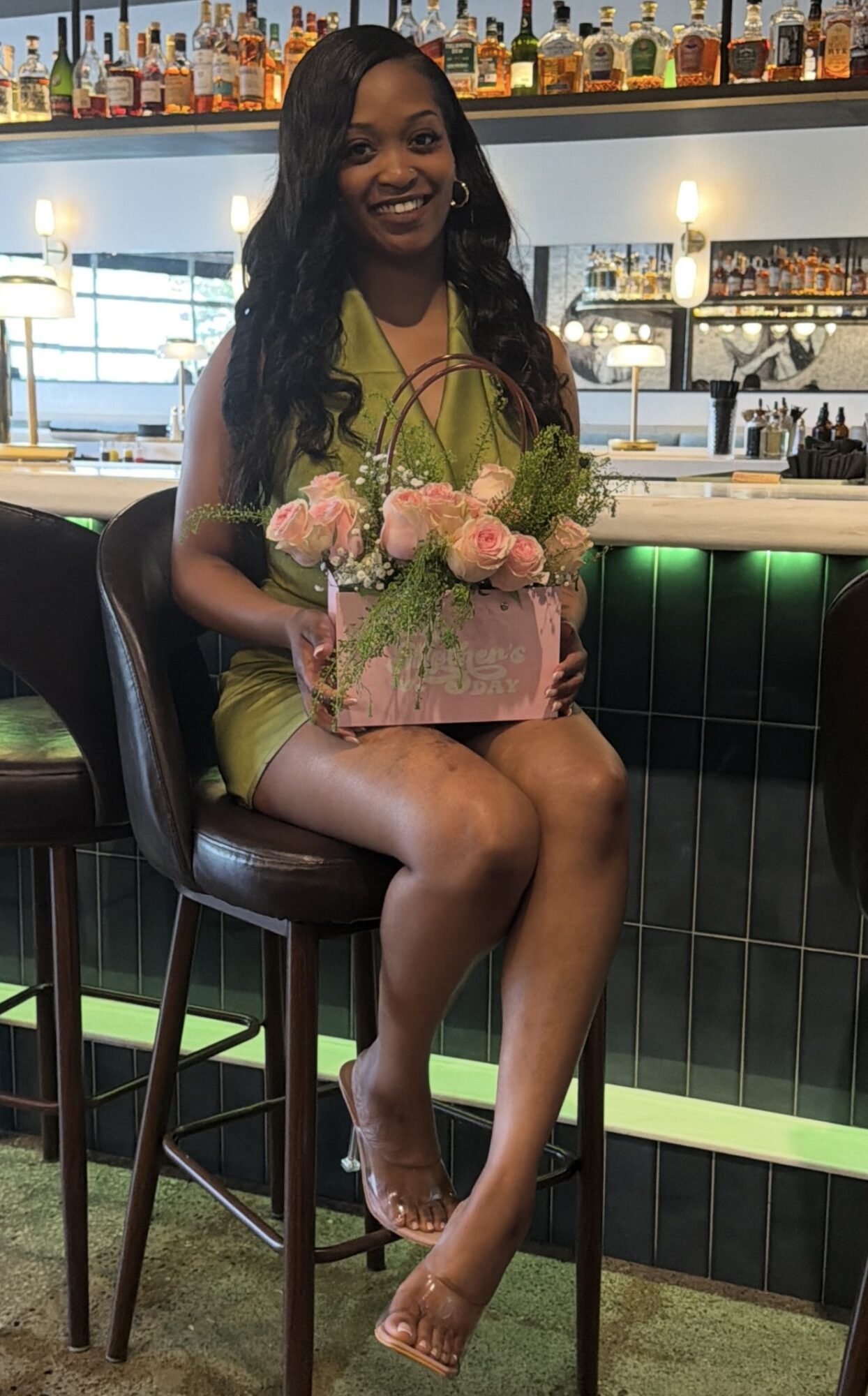 Woman sitting on bar stool holding a pink flower arrangement in a bar or restaurant setting.