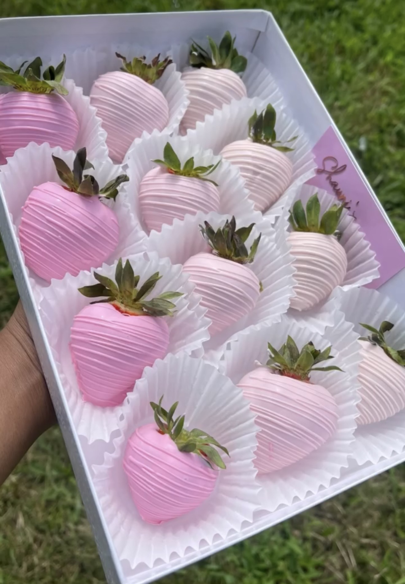 Tray of pink and white chocolate-covered strawberries with green leaves, arranged in white paper cups, outdoors on grass.
