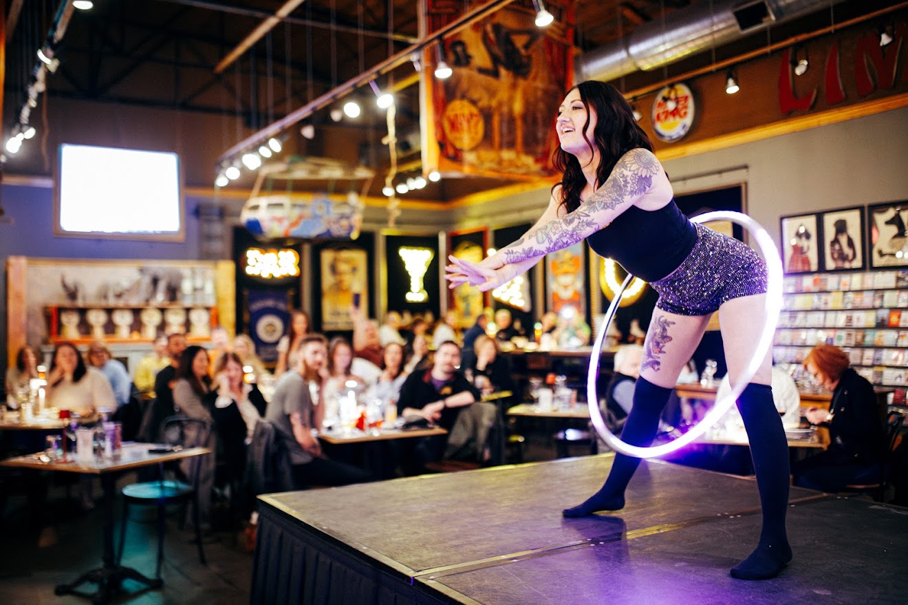 A woman performs on stage in a lively bar or restaurant with an audience watching, colorful decorations, and framed pictures on the wall.