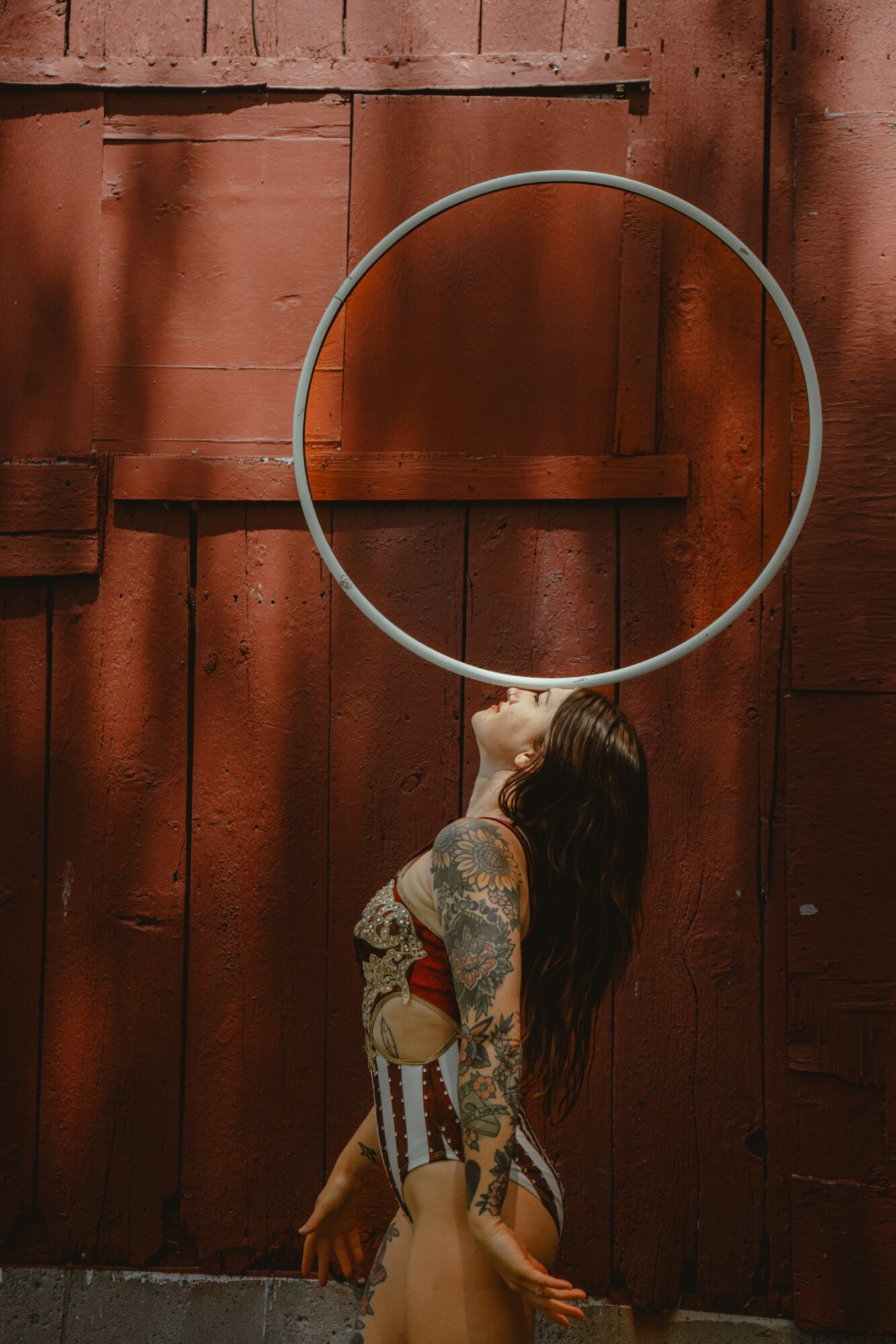 Person with long hair and tattoos stands in front of a wooden wall, holding a large metal ring overhead.