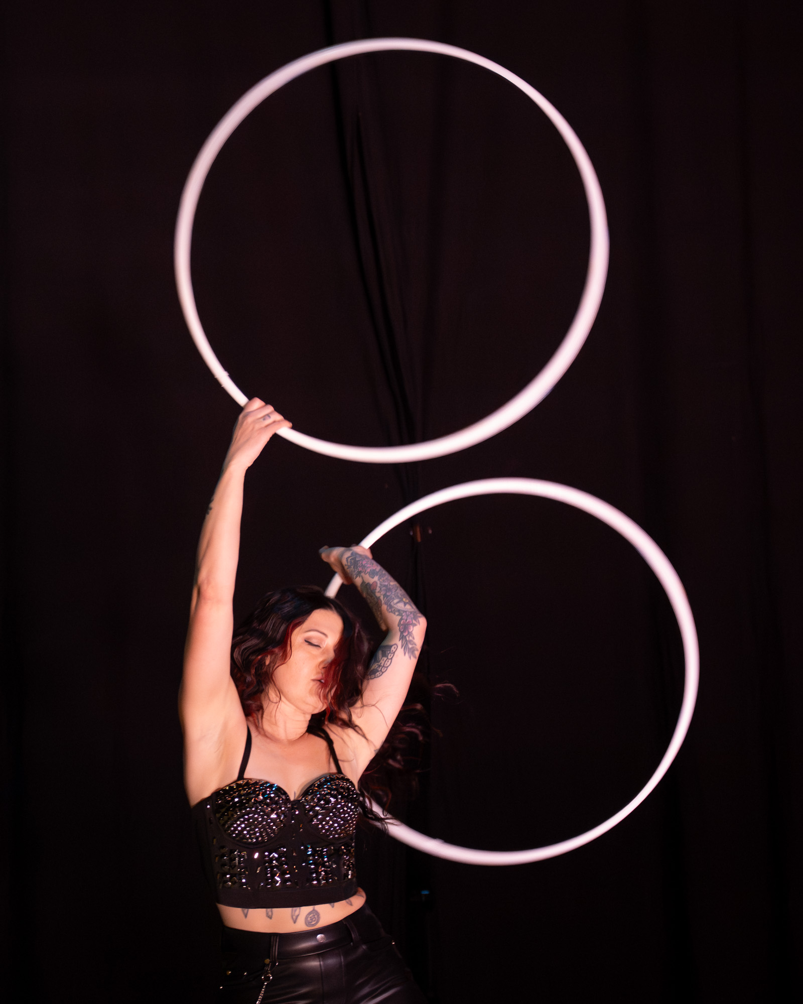 A woman performs with two large illuminated hoops against a dark background, holding one hoop above her head and the other at her side.