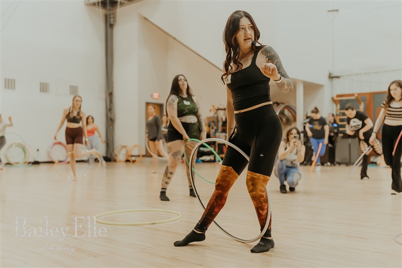 Woman in black dress holding a hula hoop in a gymnasium with other people in the background.