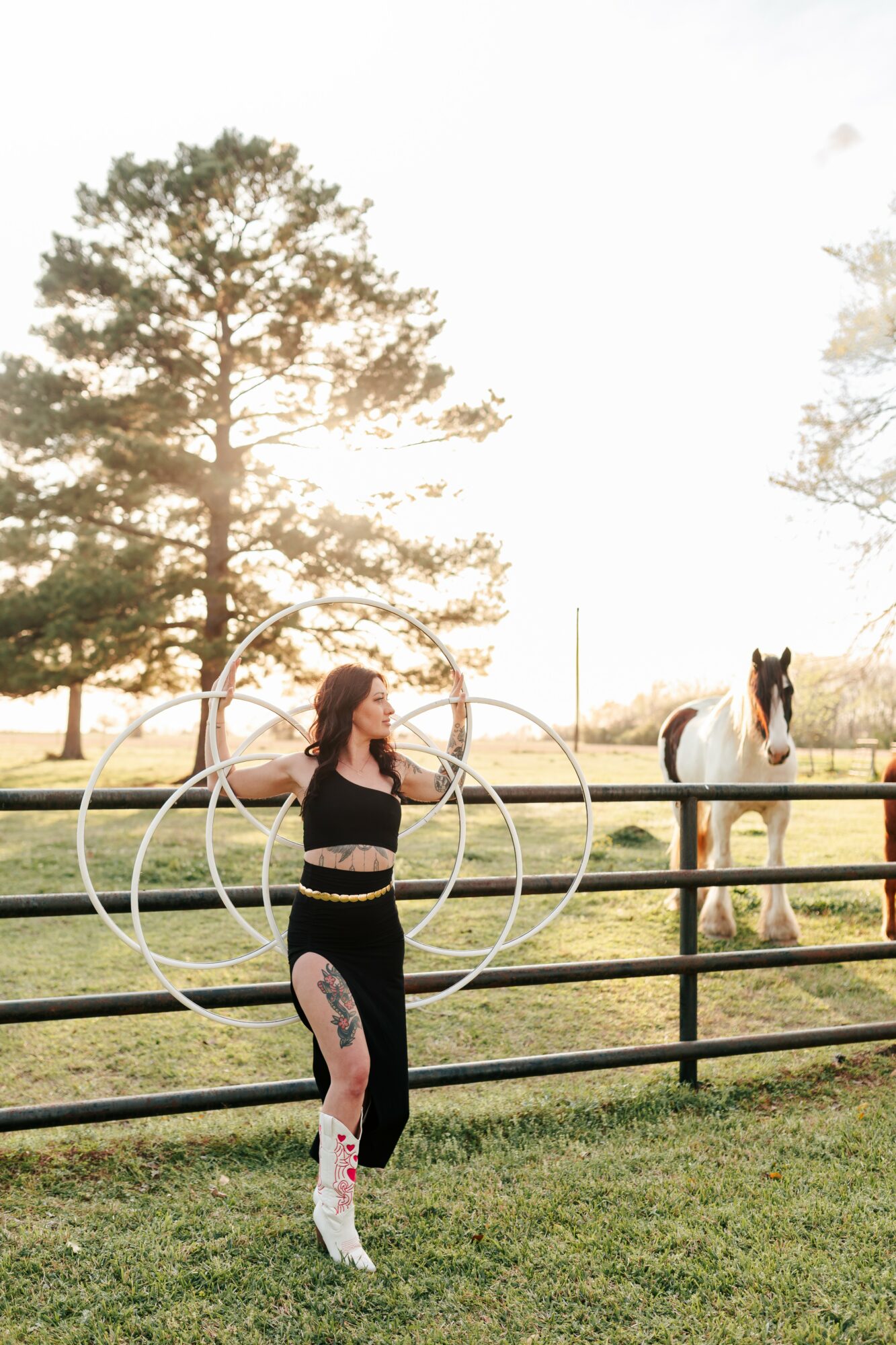 Woman standing near a fence with a horse in the background, trees, and a bright sky.