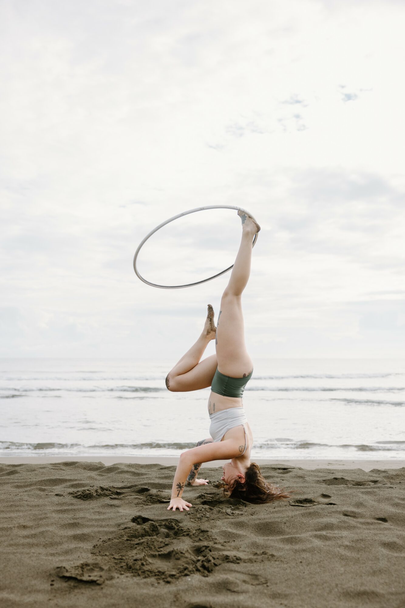 Person performing a headstand on the beach, holding a hula hoop above their foot, with water and sky in background.