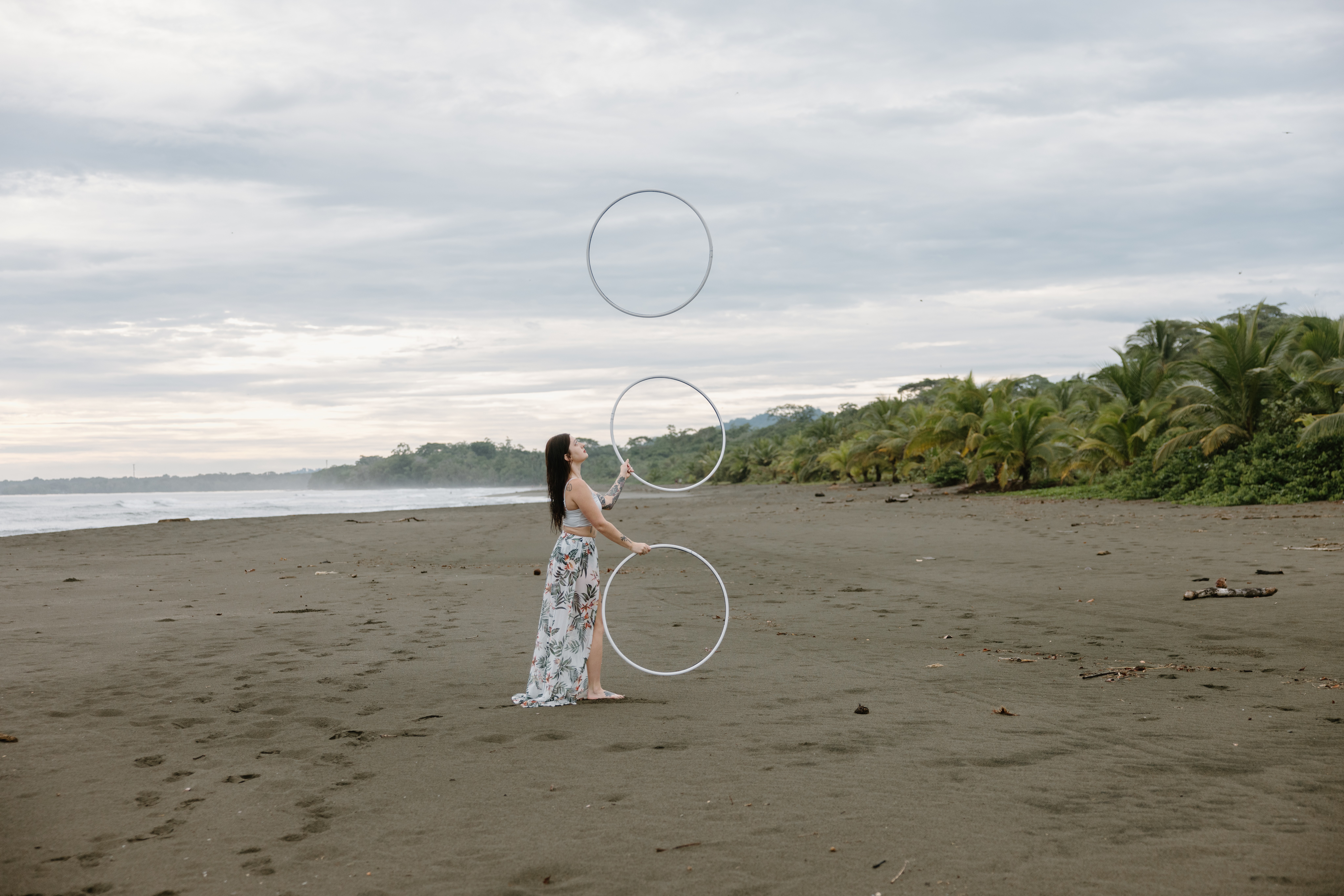 Woman standing on a beach with palm trees in the background, looking at the sky, overcast sky, sandy shore, 20 words