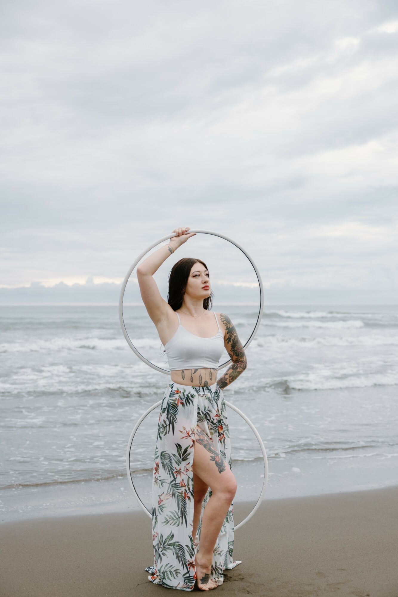 Woman standing on beach holding two hoops, wearing a white top and floral skirt, with ocean and cloudy sky in background.