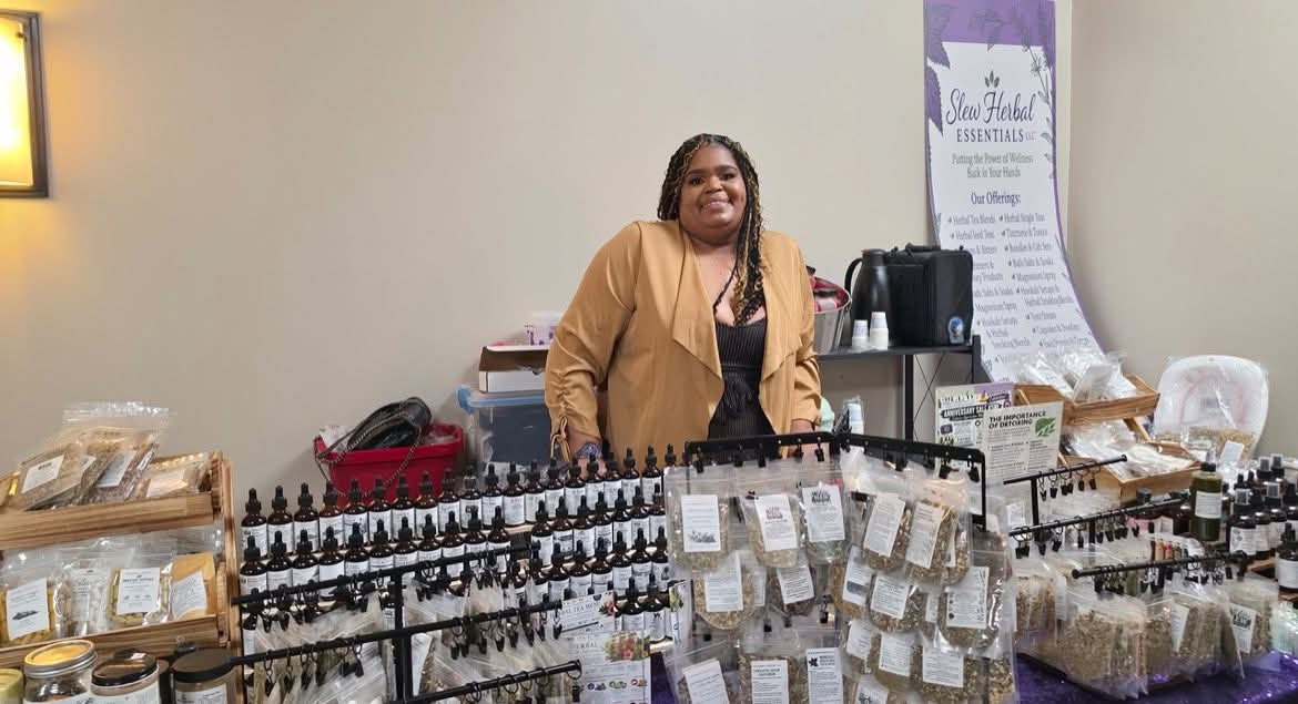 Smiling woman standing behind a table with various small bottles and products at an indoor market or event.