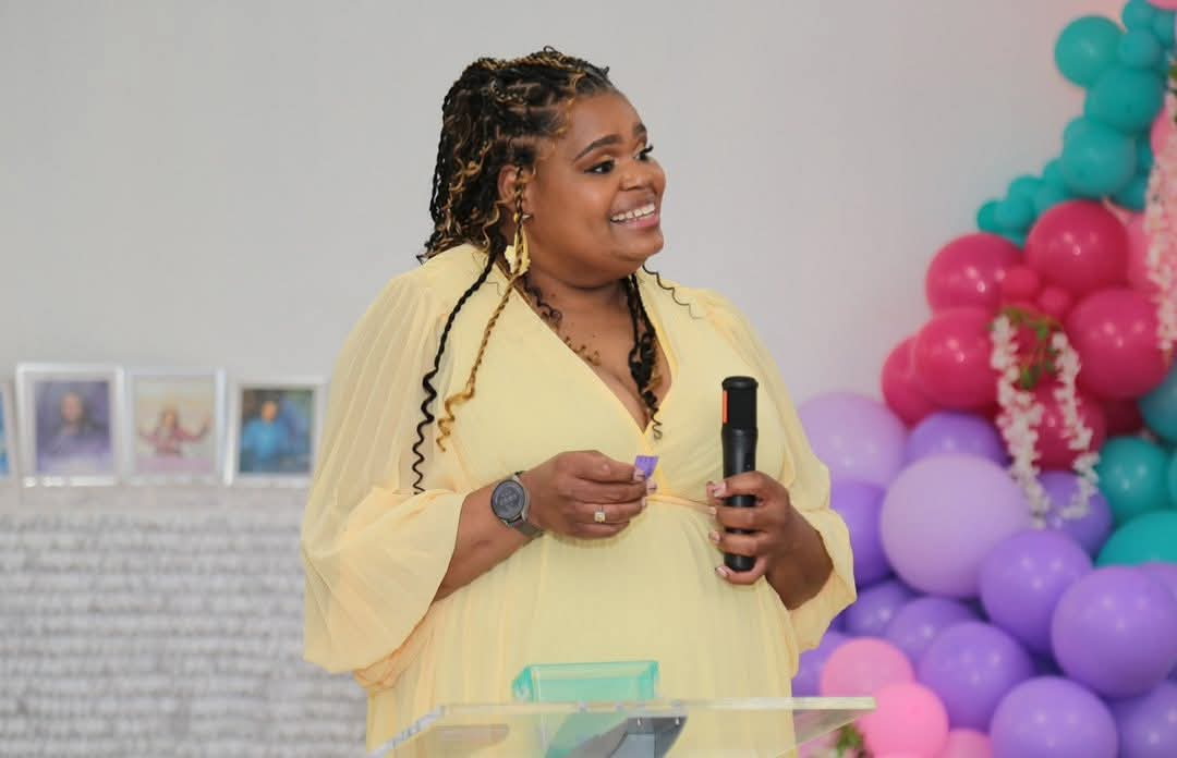 Woman speaking into a microphone at a celebration with colorful balloons in the background.