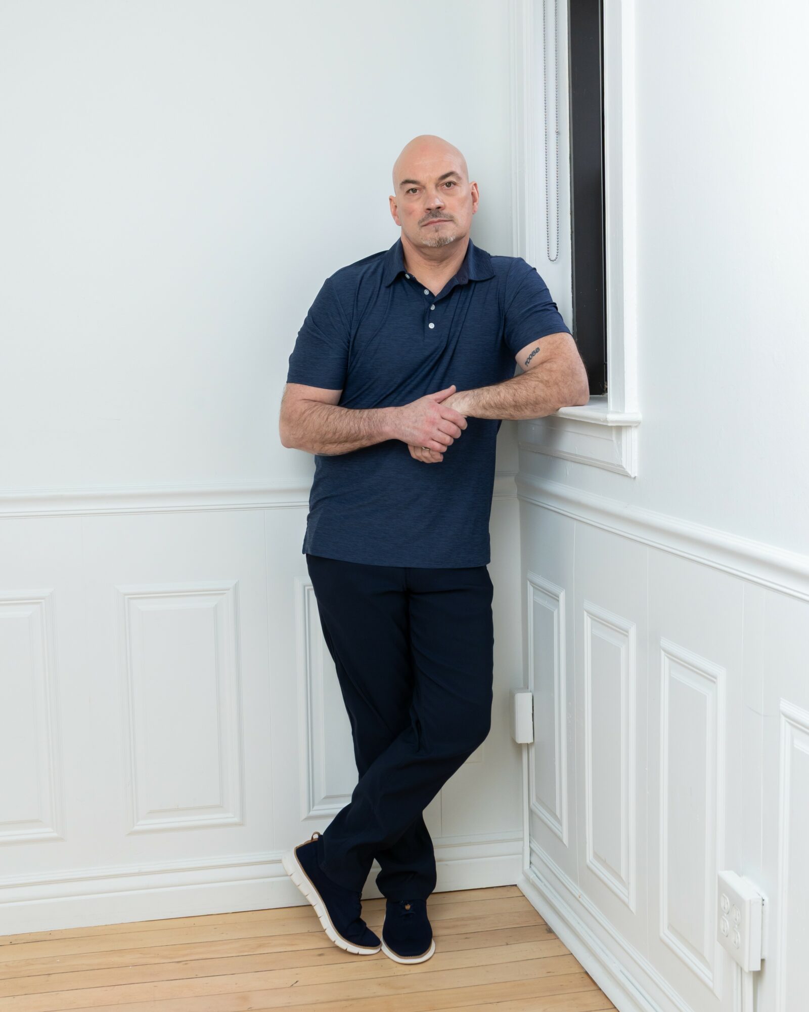 Man leaning against wall near window, arms crossed, in a room with white walls and wooden floor.