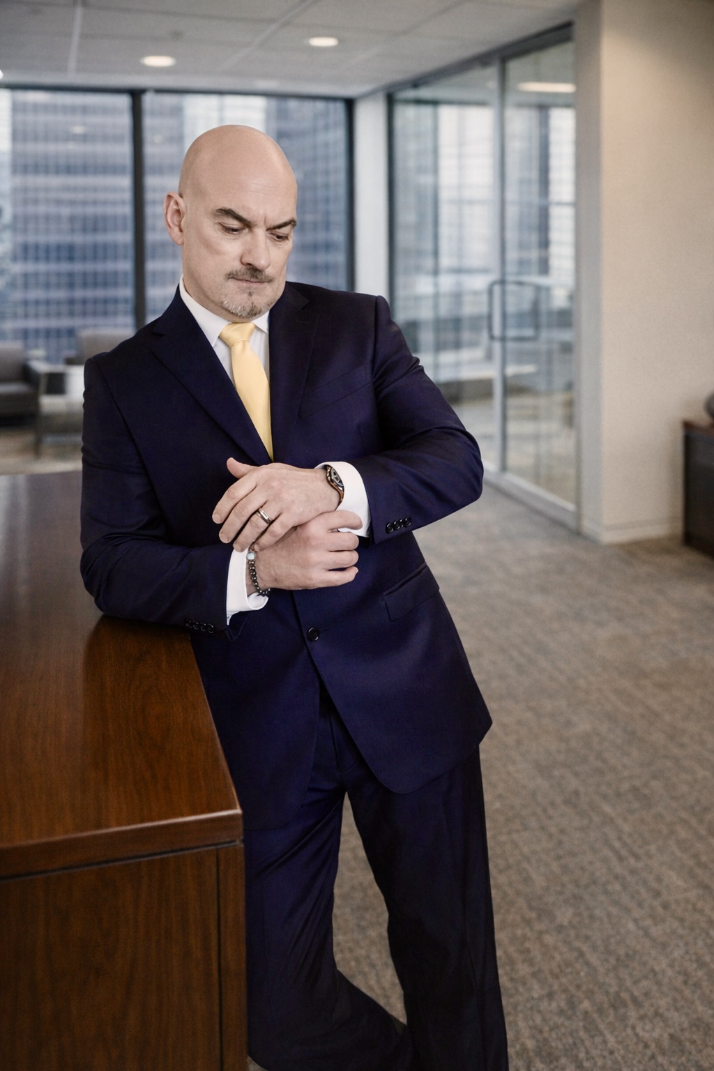 Bald man in a dark suit and yellow tie looking at his watch in a modern office with large windows.