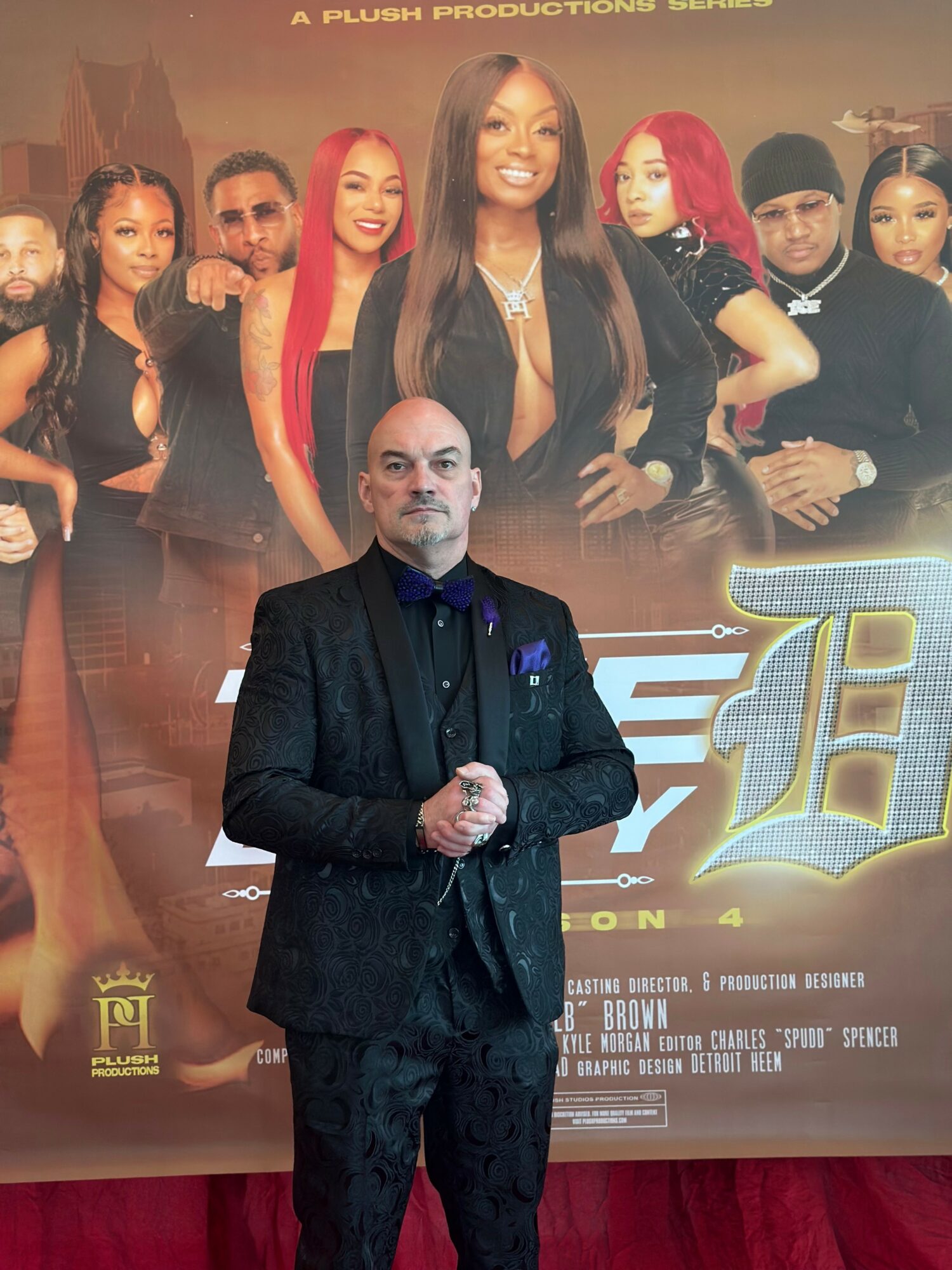 Man in a dark suit standing in front of a group photo of diverse women, some with red hair, in front of a promotional poster.
