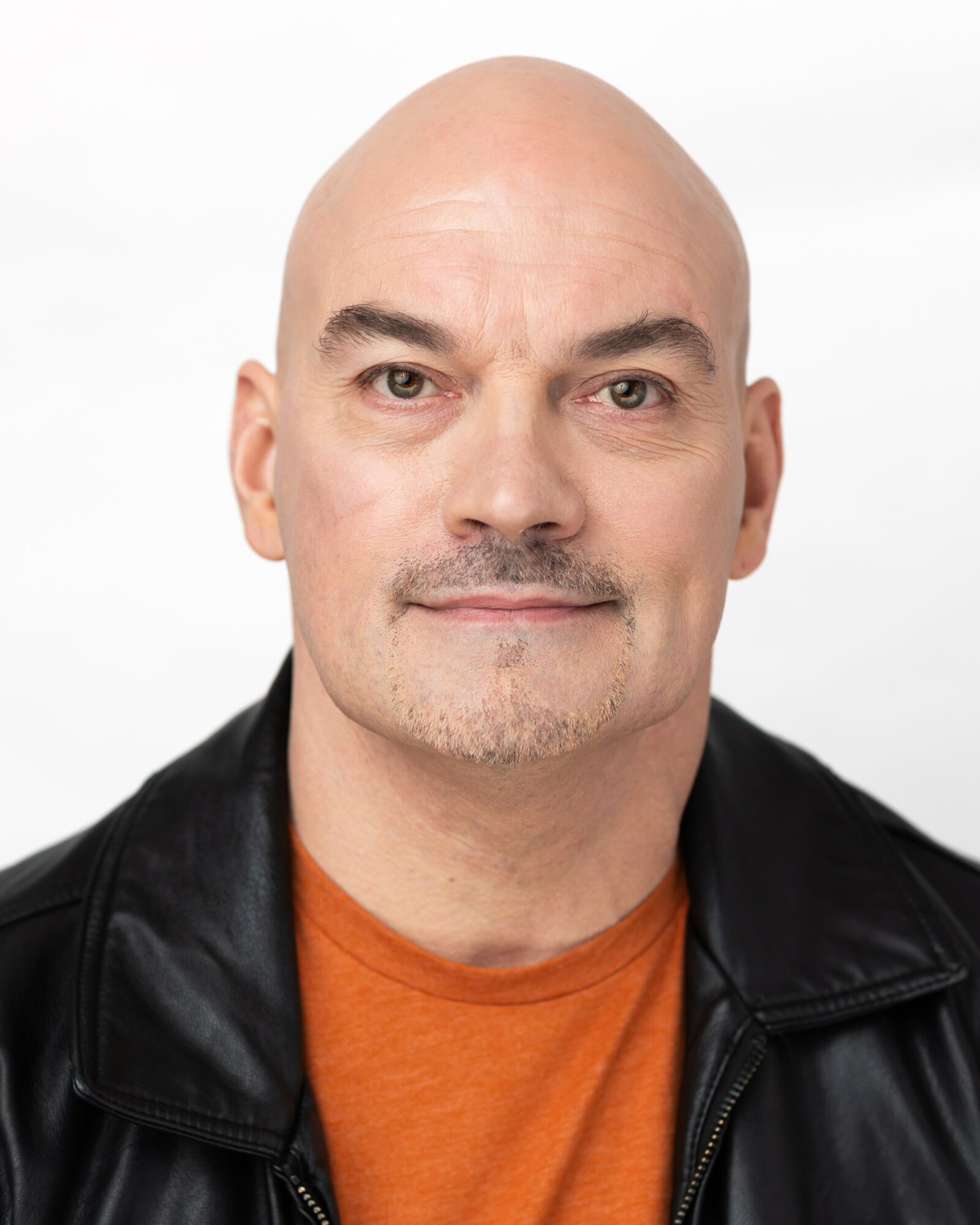 Bald man with a slight smile, wearing a black leather jacket and orange shirt, against a white background.