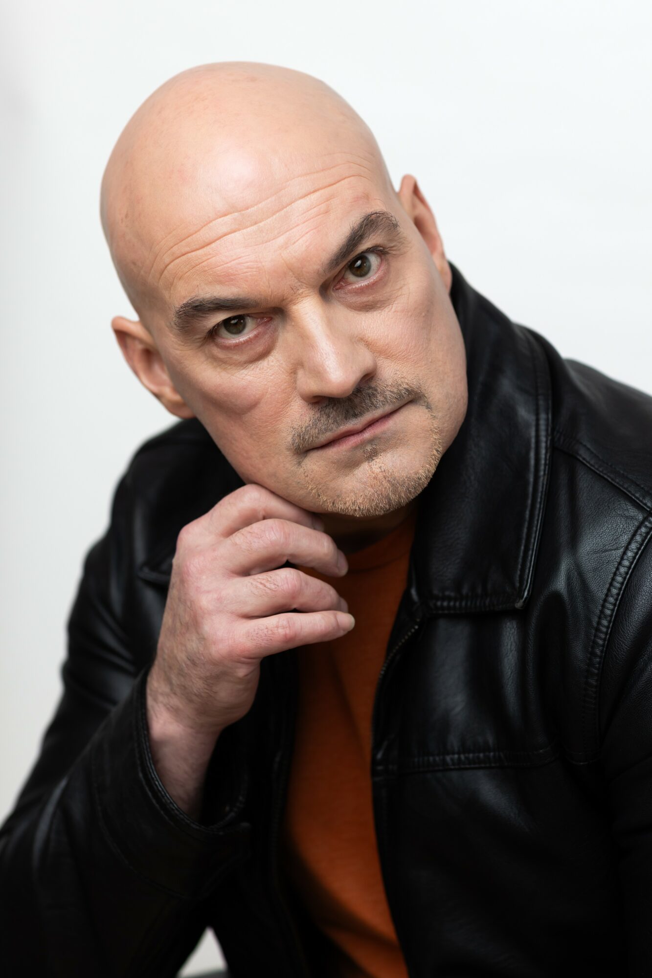 Bald man with serious expression, wearing a black leather jacket, resting his chin on his hand, against a plain white background.