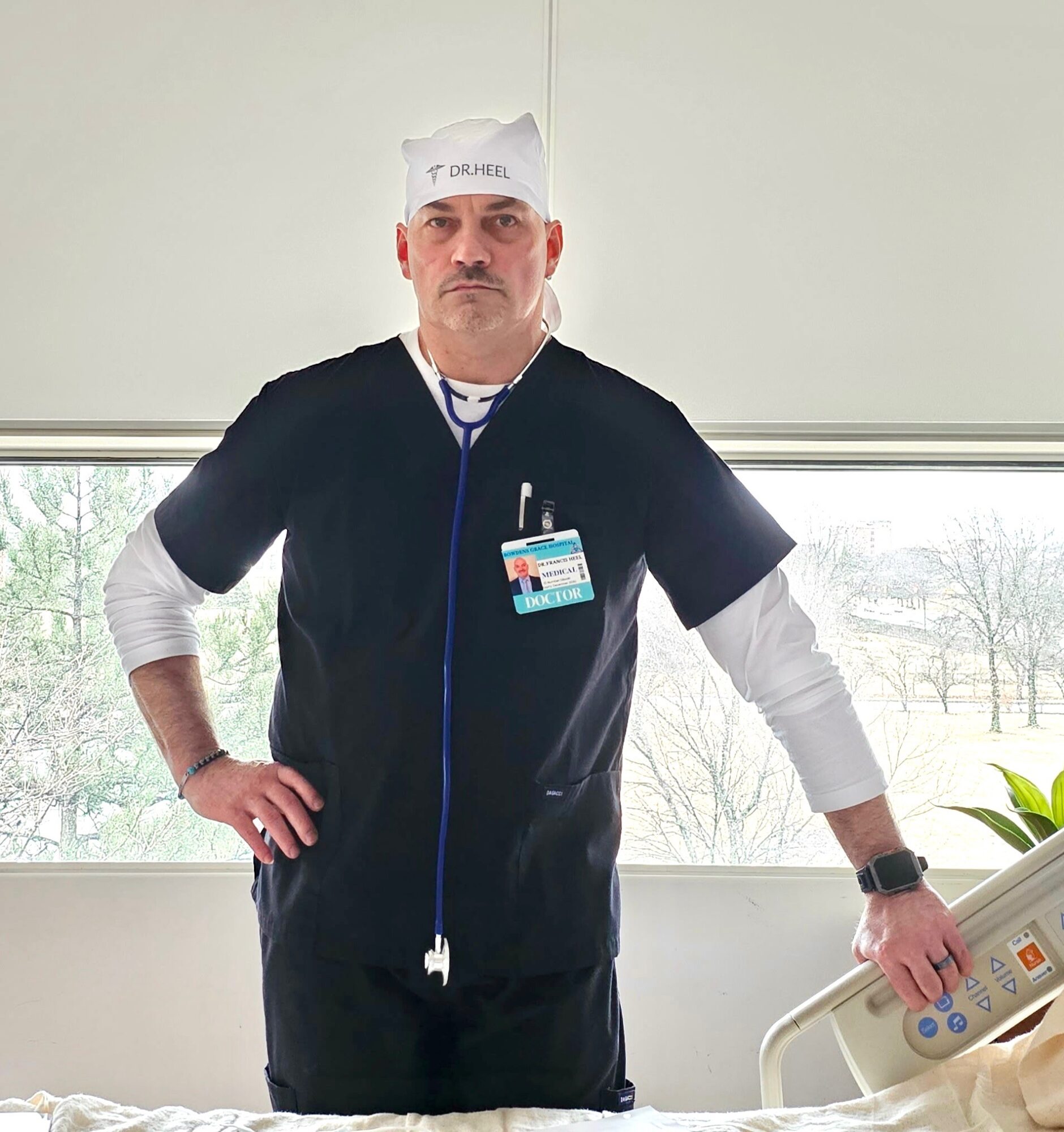 Male healthcare worker in scrubs and cap standing in a hospital room with a window and hospital bed.