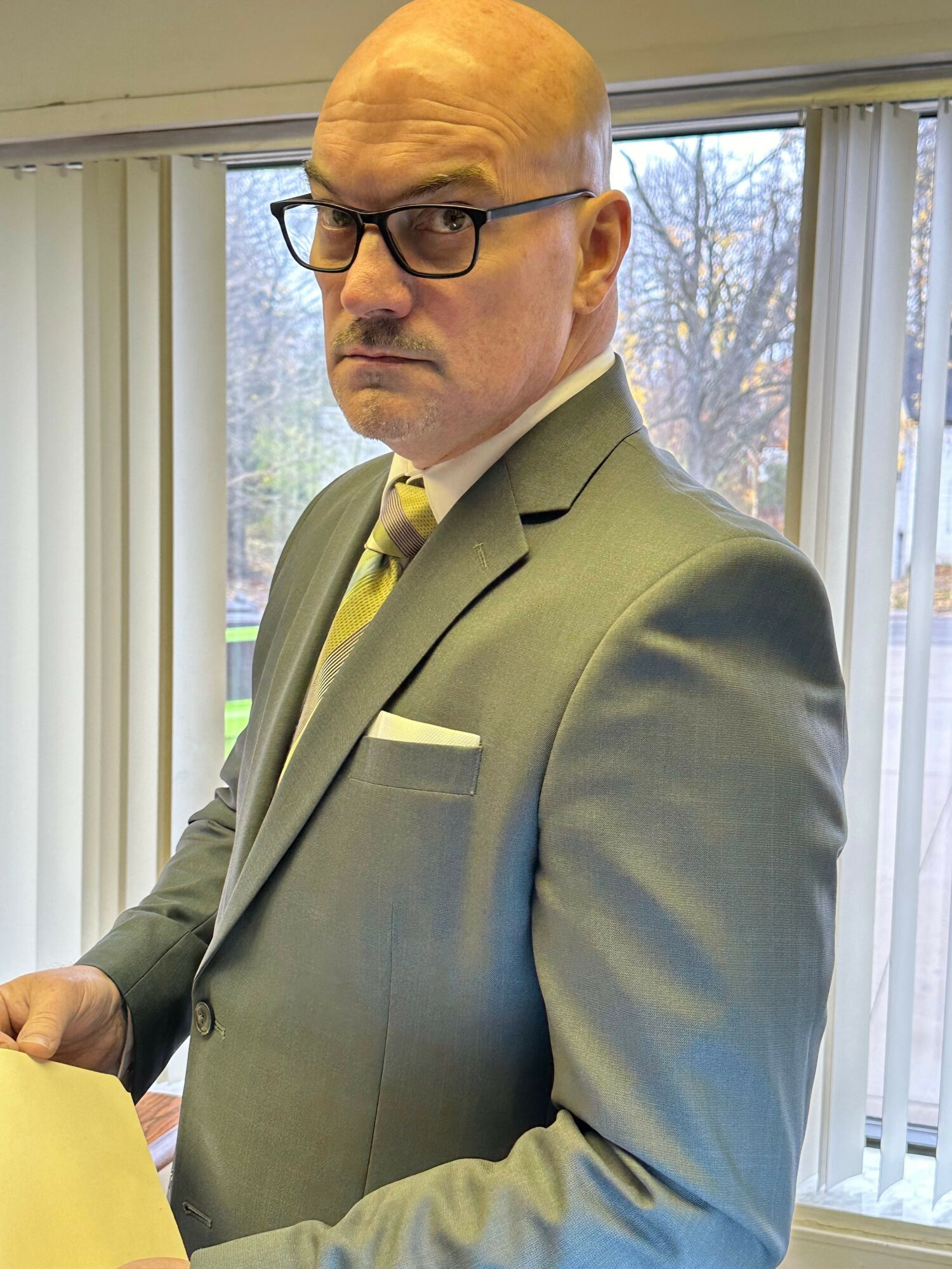 Man in a gray suit with glasses standing indoors near a window, looking to the side.
