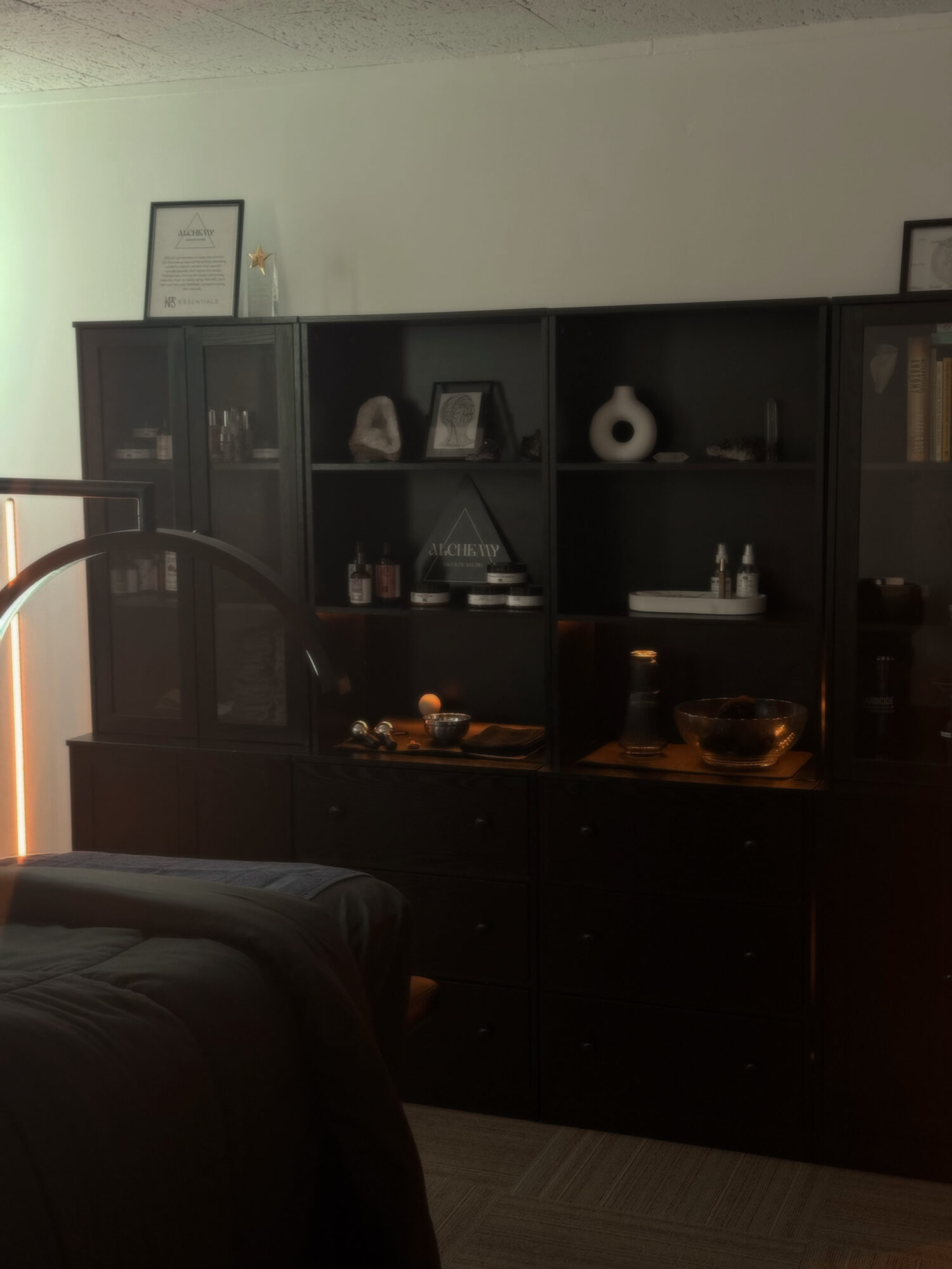 Dark cabinet with shelves holding decorative items and books, against a light-colored wall, with part of a bed in foreground.