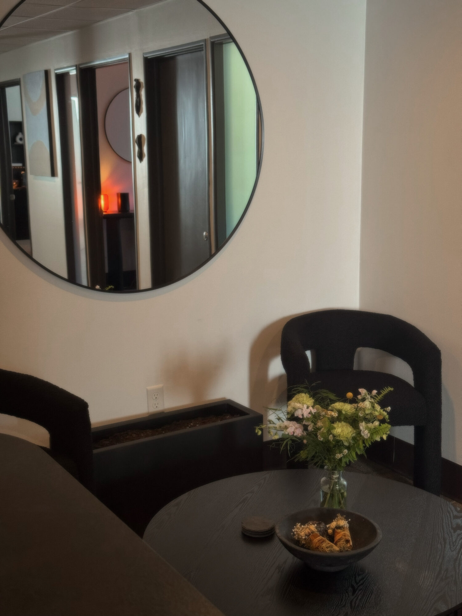 Round mirror on wall reflects hallway with doors and a lit red lamp, with a table, chairs, and a flower arrangement nearby.