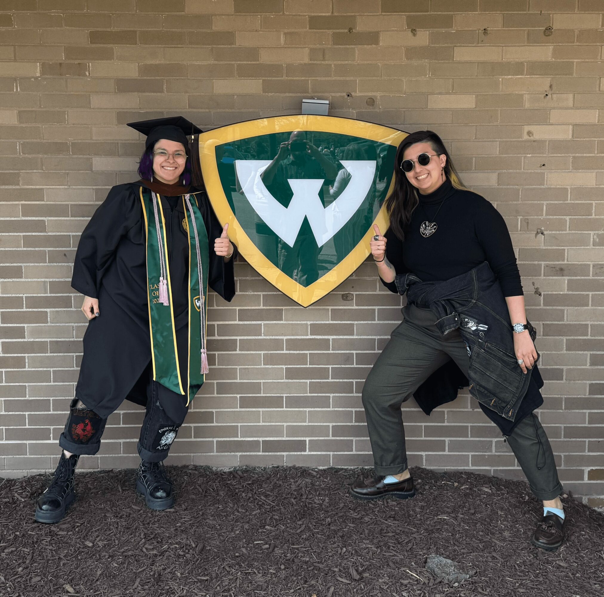 Two women stand on either side of a large green and yellow shield emblem on a brick wall, smiling and posing.