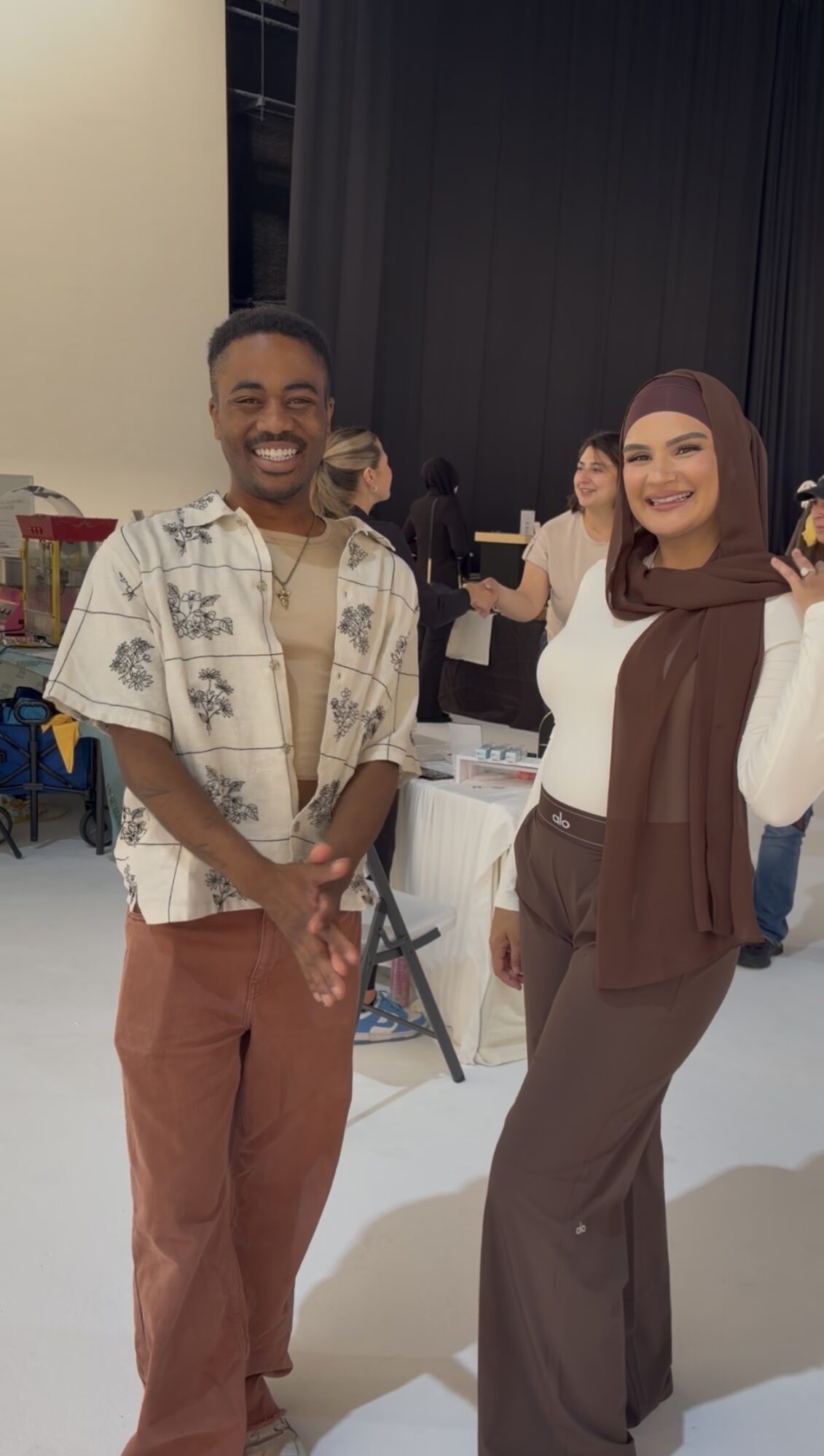 Two people smiling and standing indoors, one wearing a patterned shirt and the other in a brown vest and pants.
