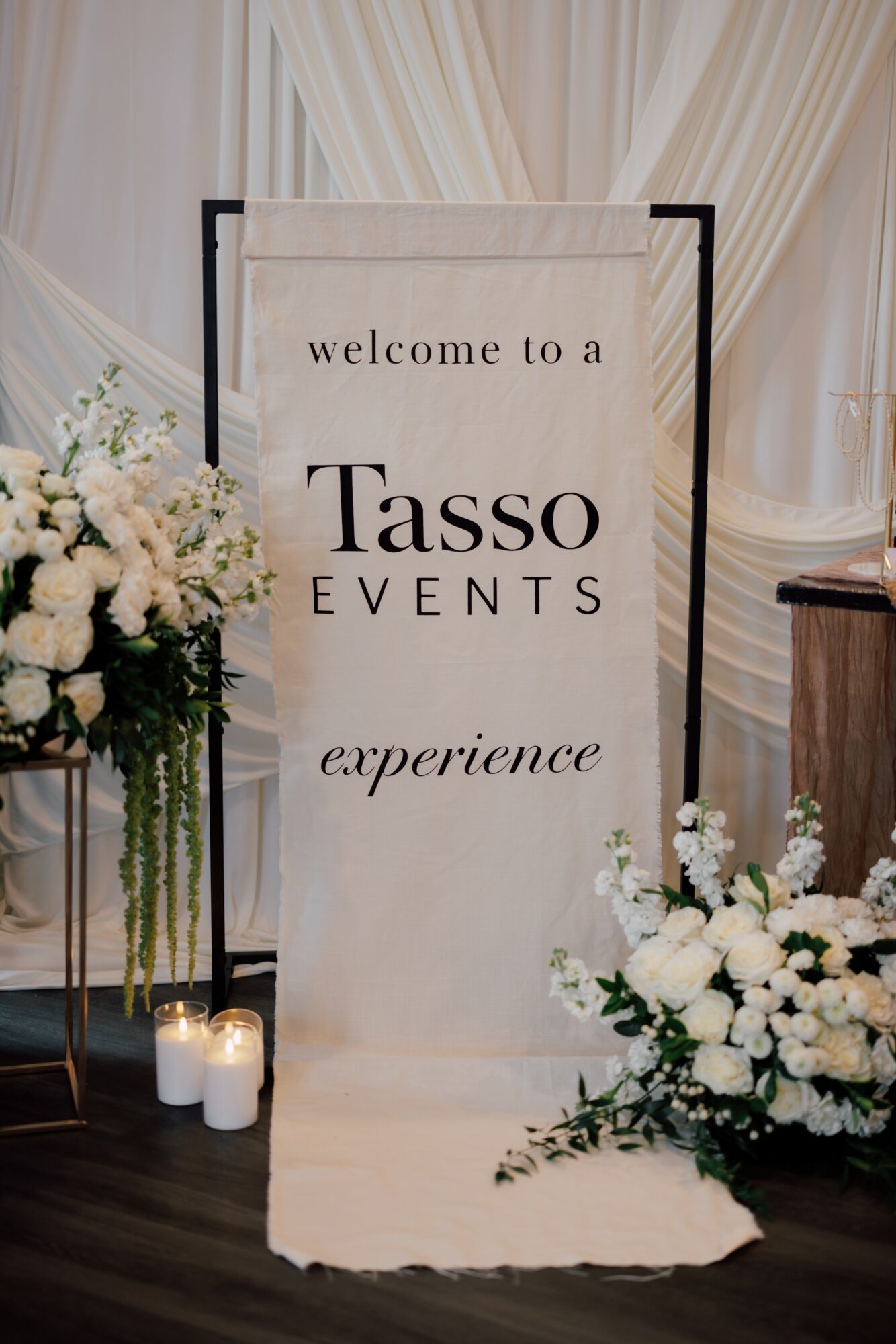 Welcome banner for Tasso Events with floral arrangements and candles nearby, set against draped curtains.