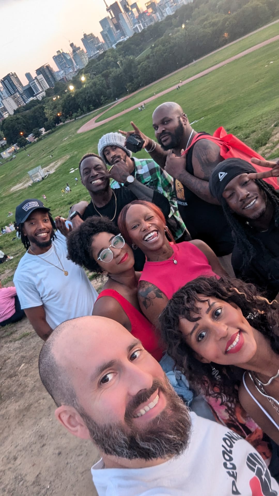 Group of nine people smiling outdoors in a park with city skyline in background.