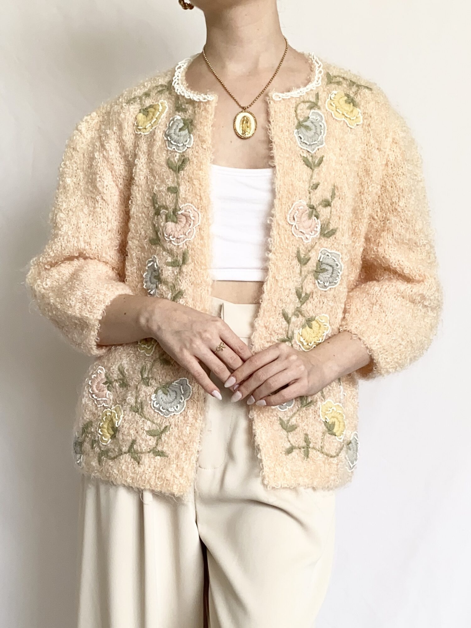 Person wearing a beige embroidered cardigan, white top, and cream pants, holding hands in front, with jewelry visible.