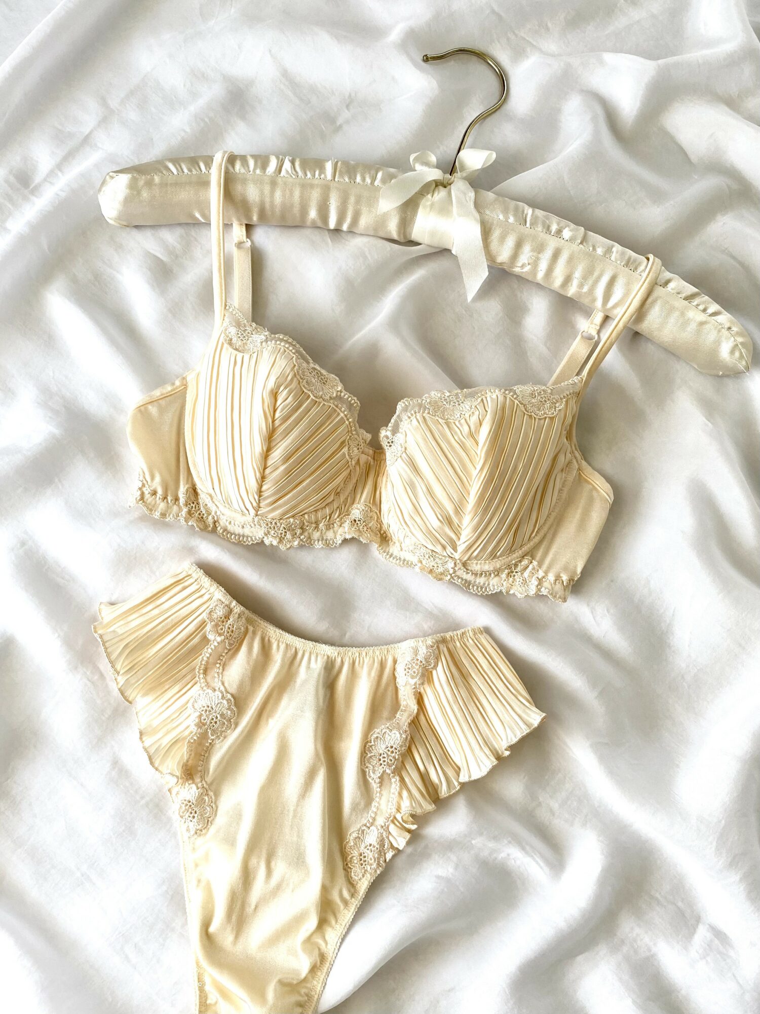 Cream-colored ruffled bra and matching panties on white fabric, with a padded hanger. The bra has vertical stripes and adjustable straps.