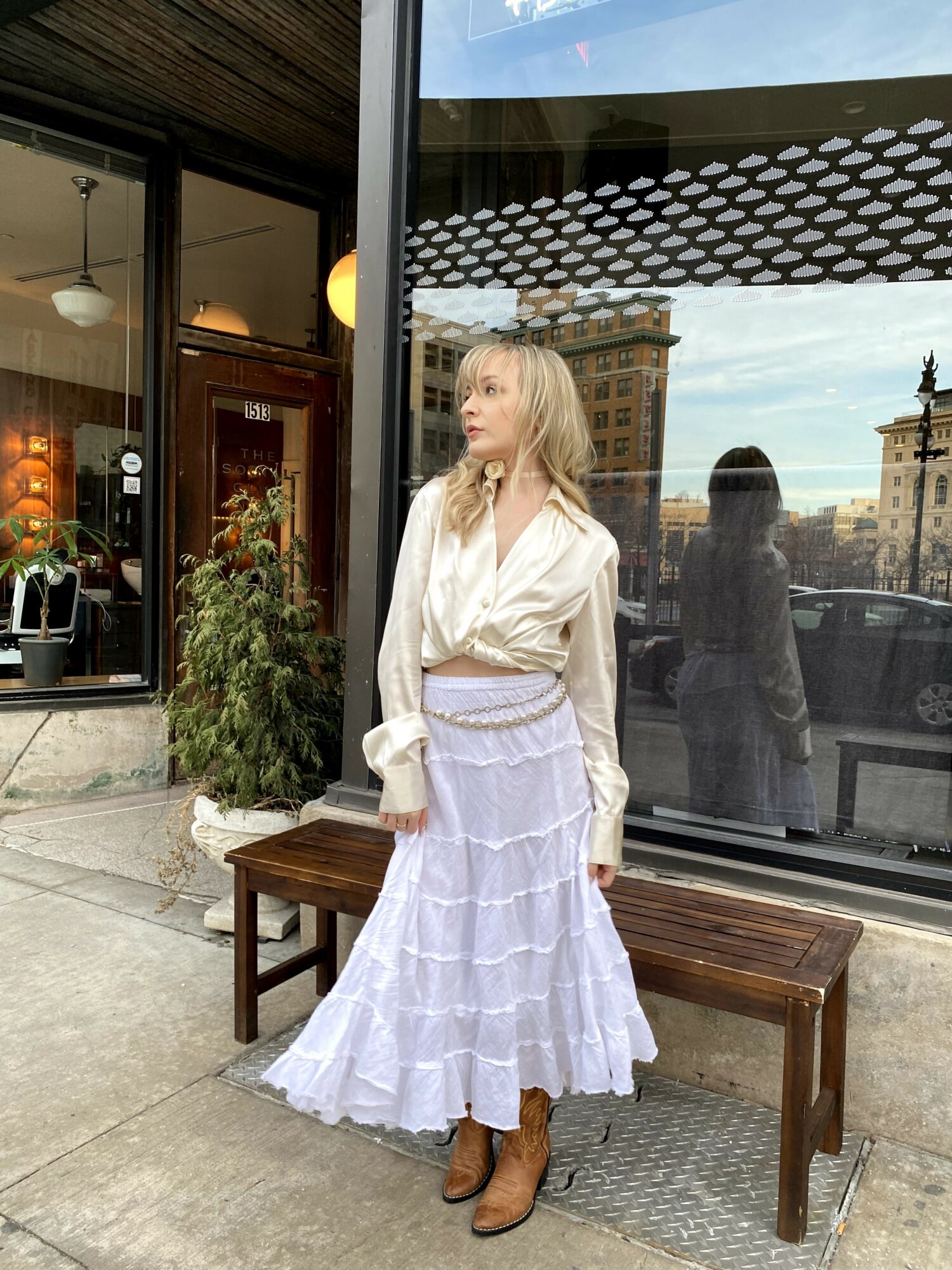 Woman with blonde hair in a white blouse and tiered skirt stands outside a building with large windows, cityscape background.