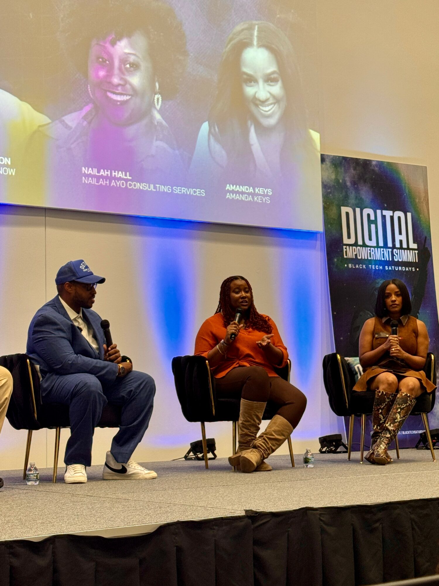 Three people seated on stage in front of a large screen displaying two women smiling. The stage has a blue background and a sign reading 'DIGITAL'.