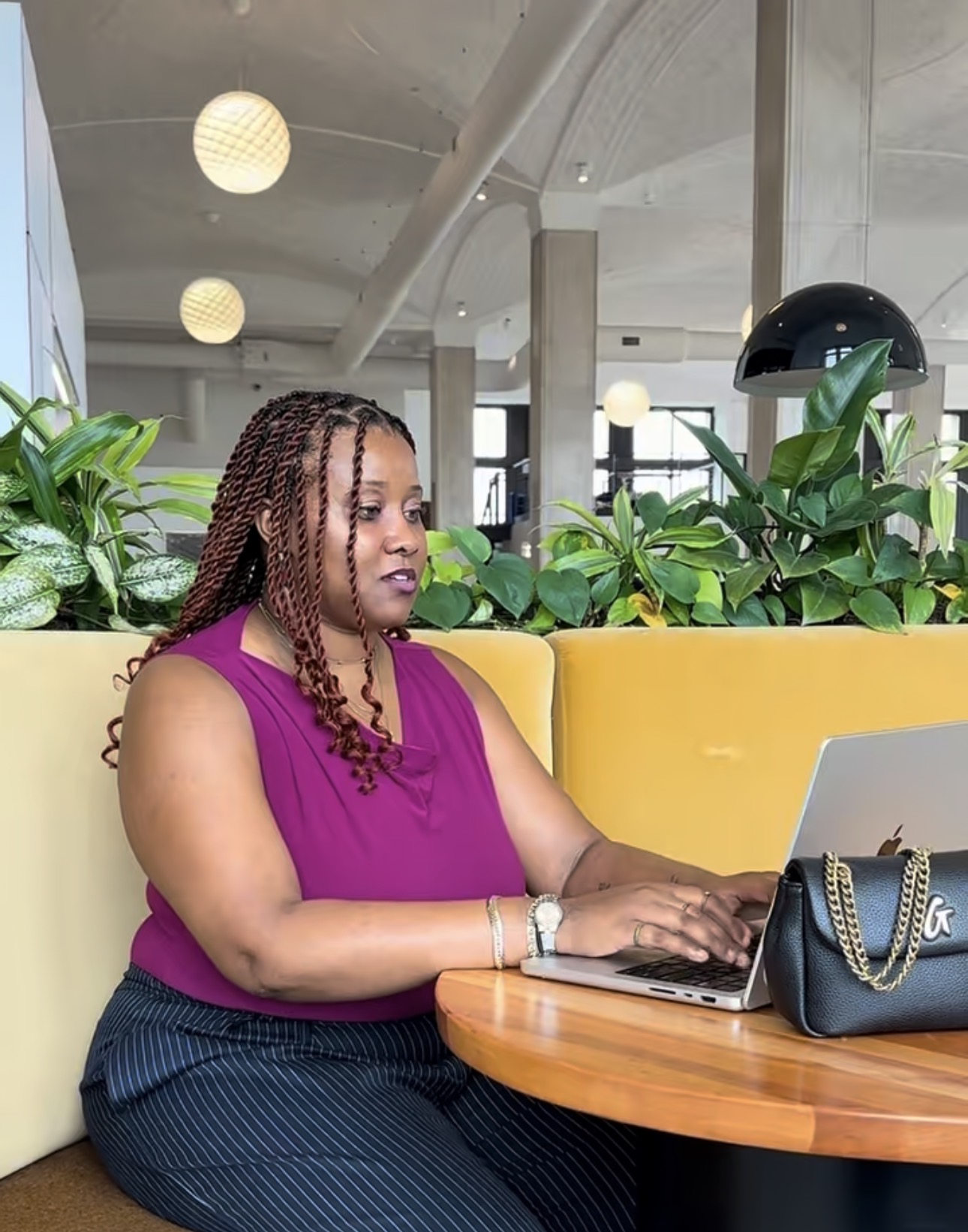 Woman with braided hair working on a laptop at a round table in a modern indoor space with plants and yellow seating.