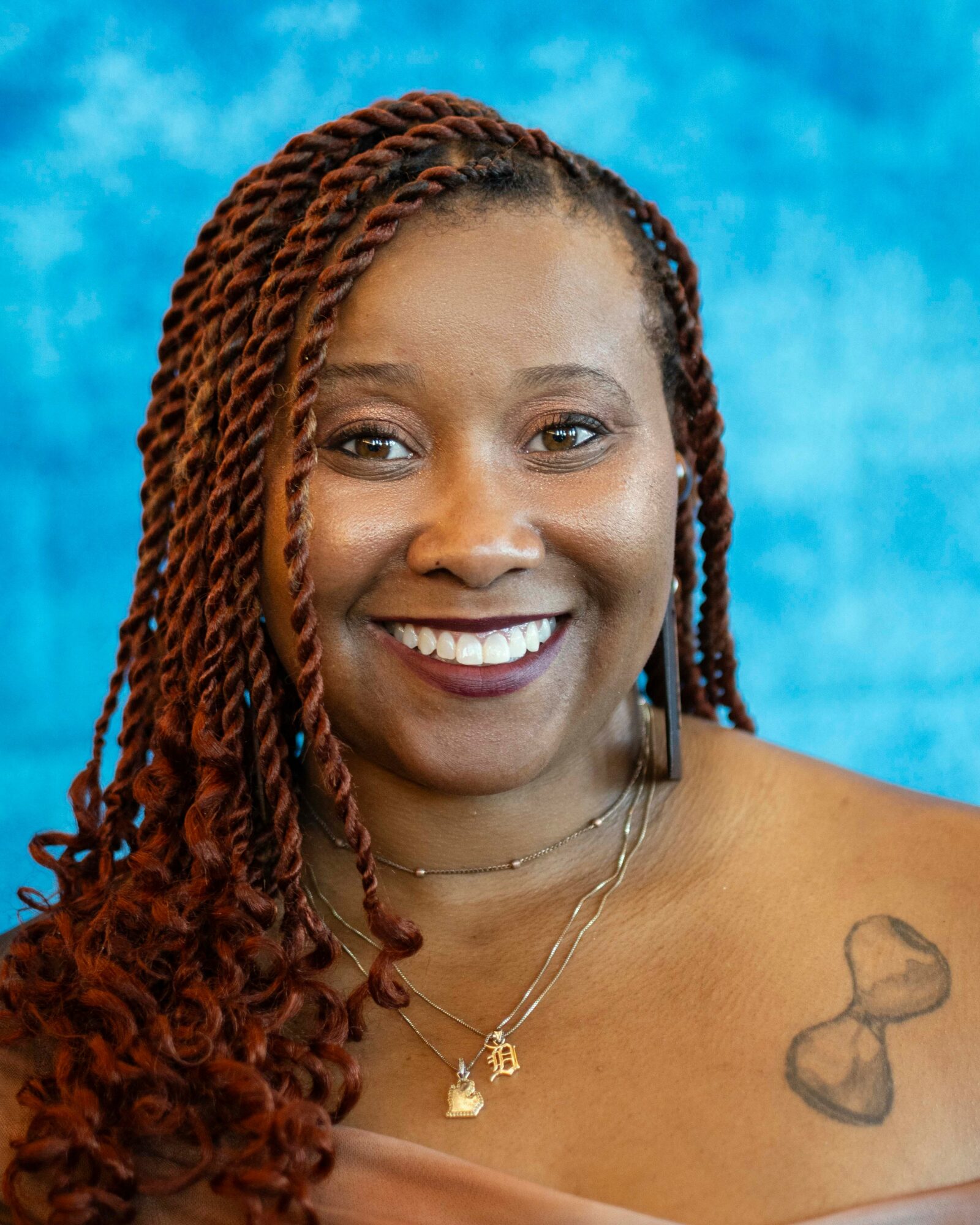 Smiling woman with braided hair, wearing necklaces, against a blue background.