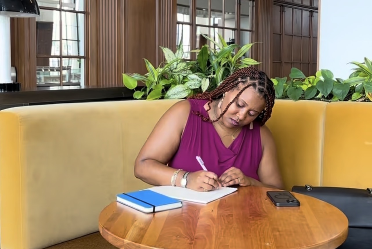 Woman with braided hair writing in a notebook at a round wooden table, with a yellow booth and green plants behind her.
