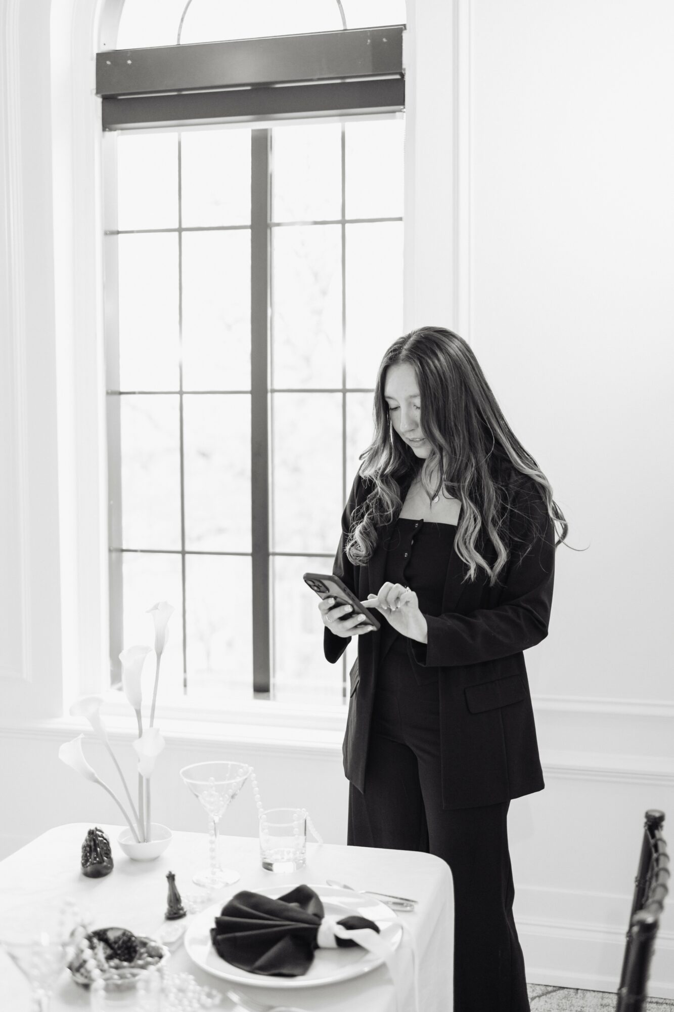 Woman with long hair looking at her phone indoors near a large window, table with items in front of her.